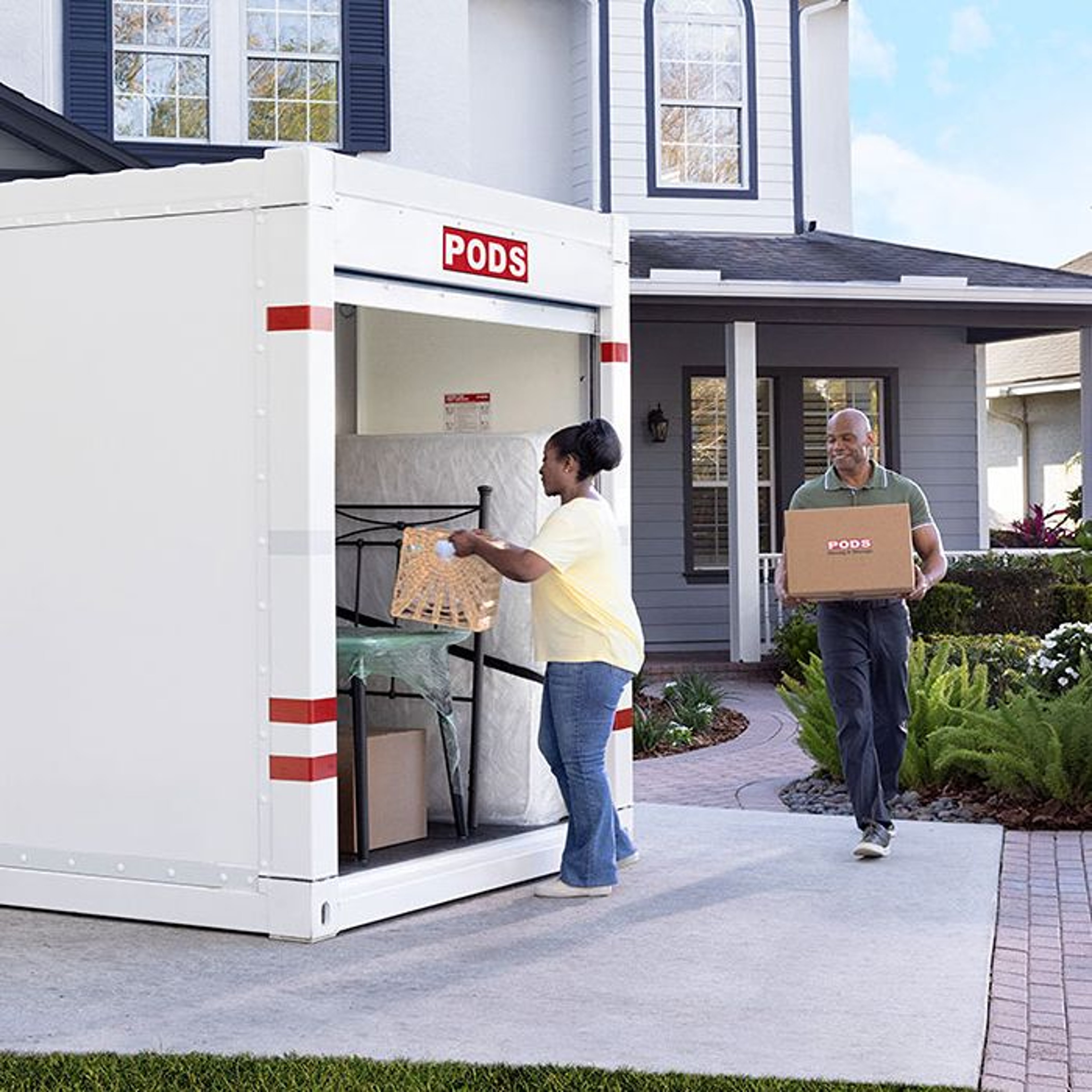 A woman loads a woven basket into a PODS storage container while a man carries a box labeled "PODS" nearby, in front of a house with a landscaped yard.