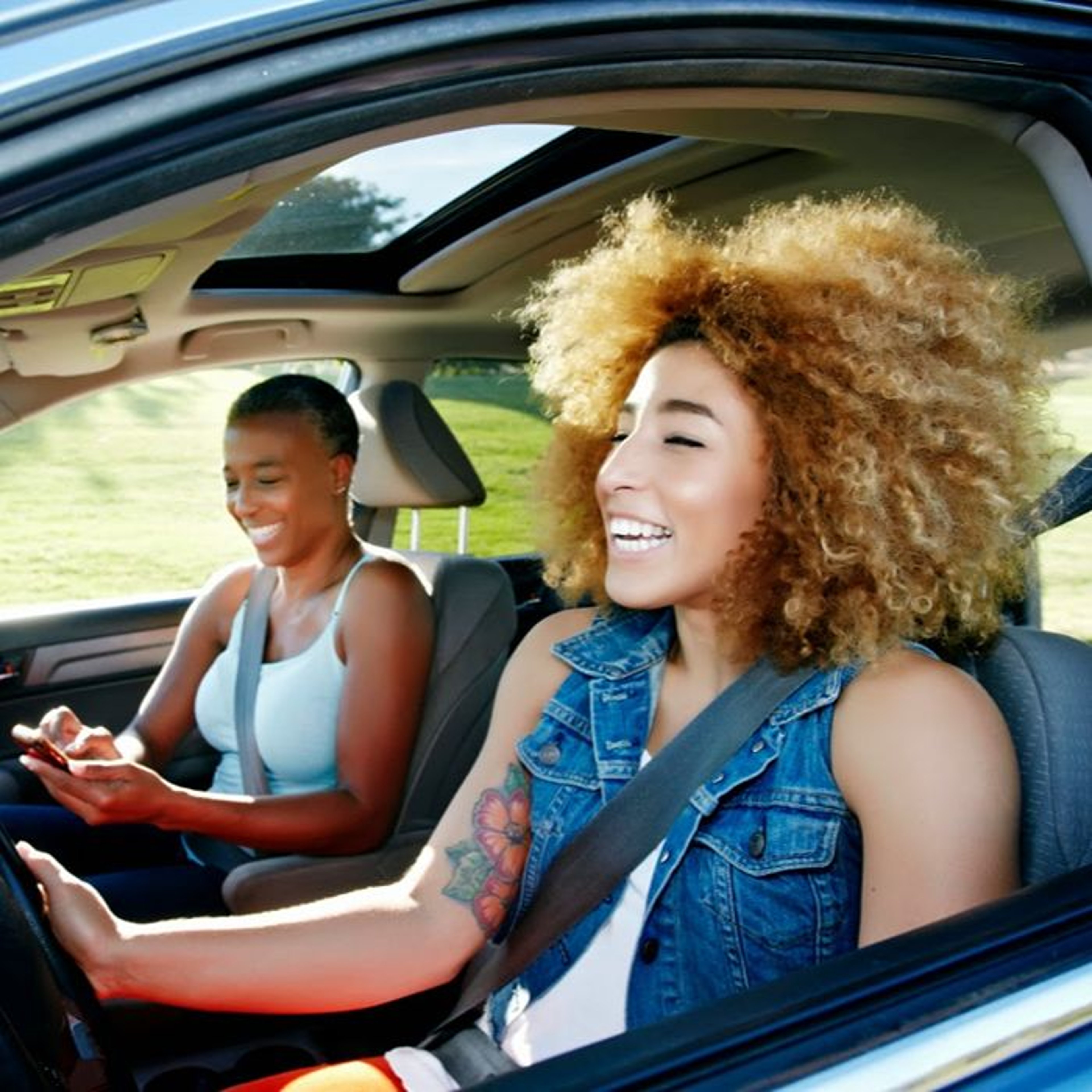 Two young women inside a car, one driving with a broad smile and curly hair, the other using a smartphone, both enjoying their time together.