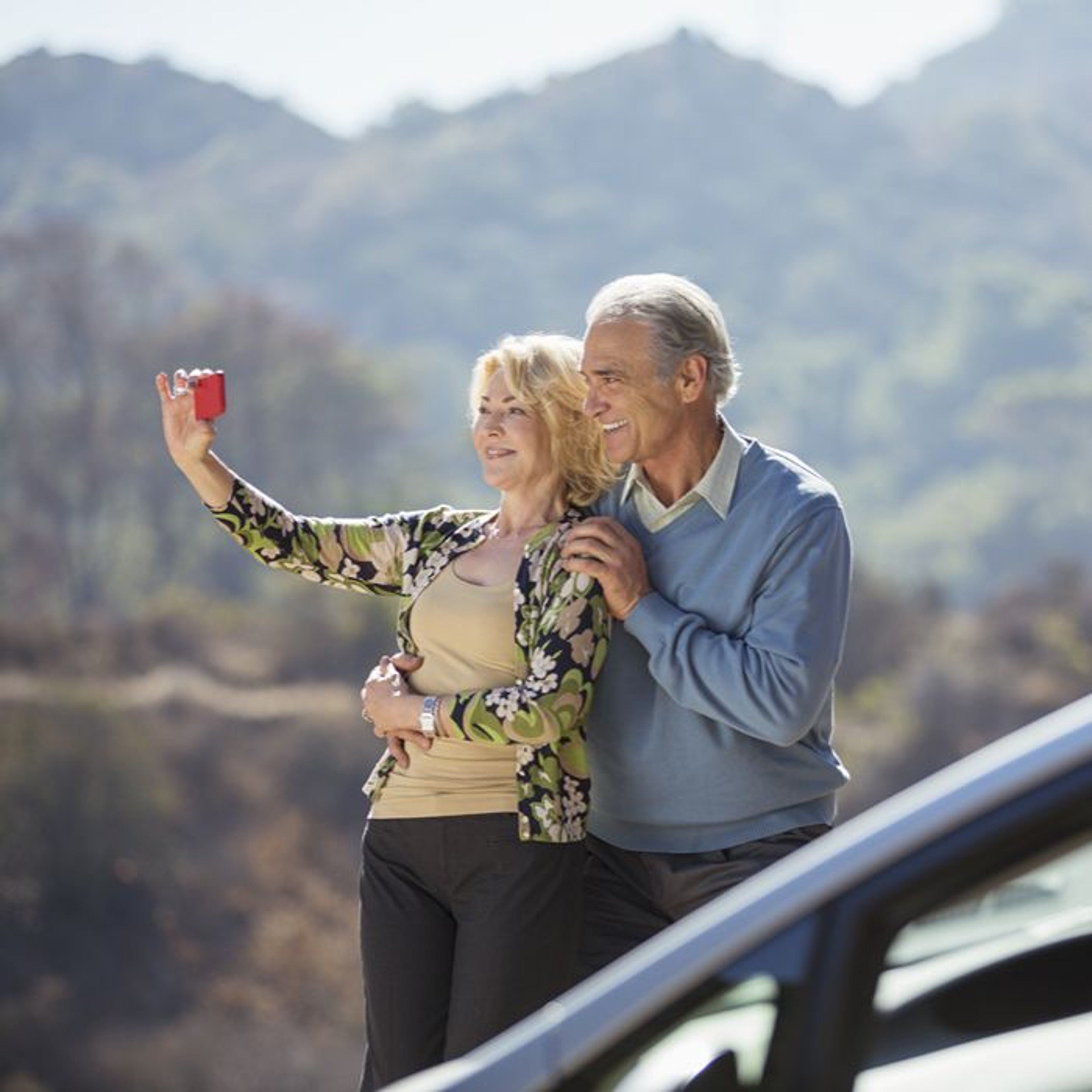 A smiling couple takes a selfie outdoors, surrounded by hills and trees. The man stands behind the woman with a gentle embrace.