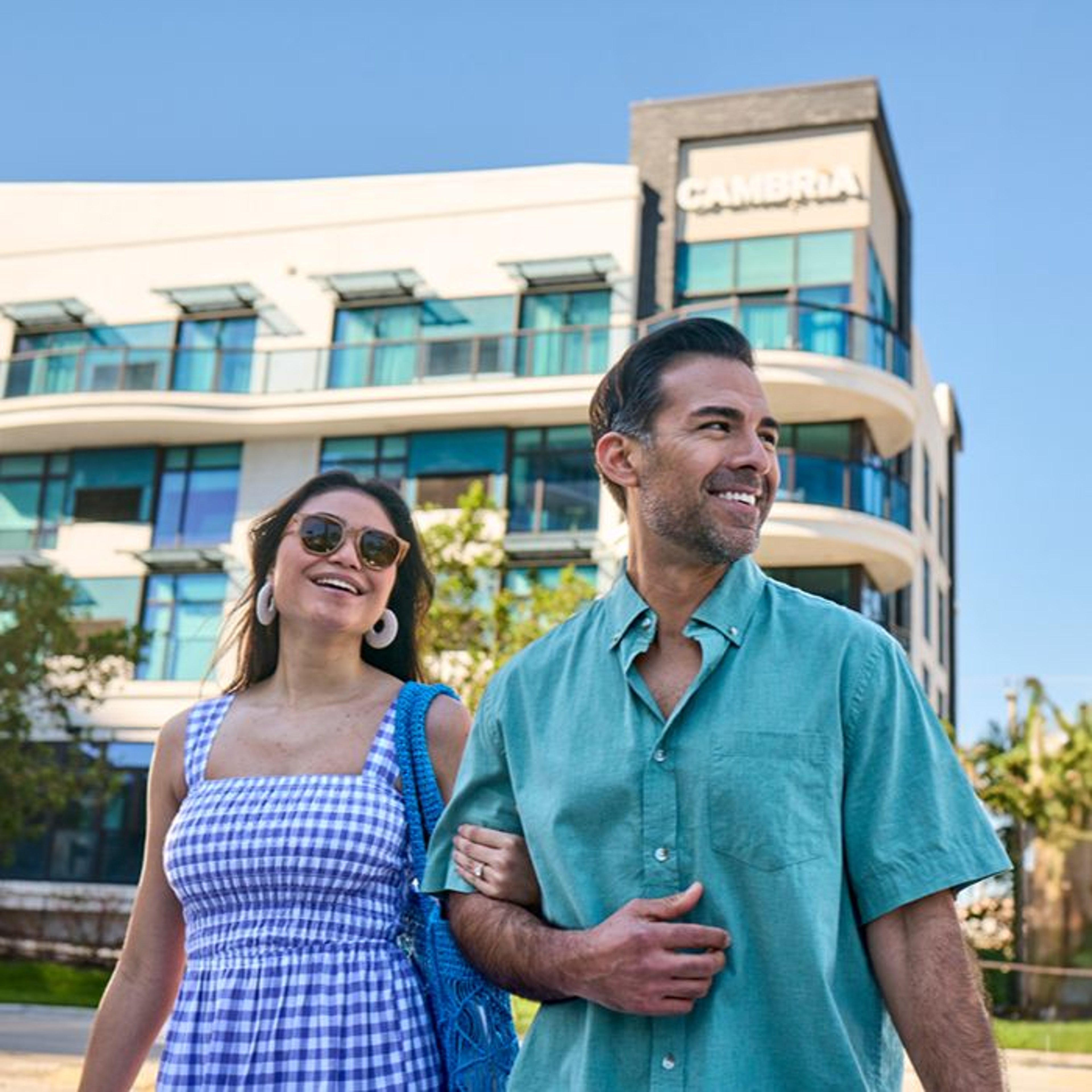 A smiling couple walks outside a modern hotel, enjoying a sunny day. The woman wears sunglasses and a blue dress.