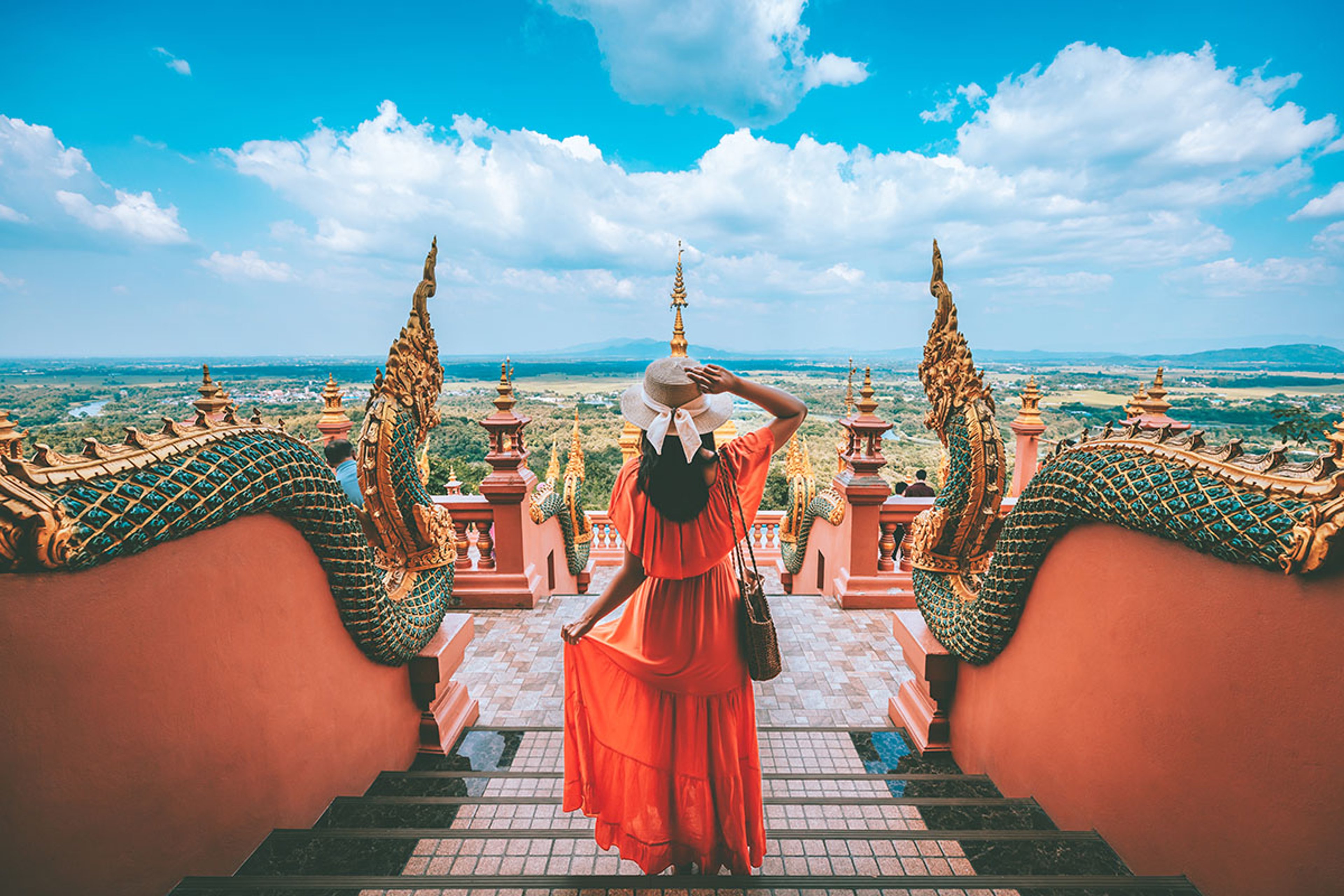 A woman in a red dress and sunhat stands on a temple staircase between ornate serpent railings, looking out over a wide landscape under a blue sky with clouds.