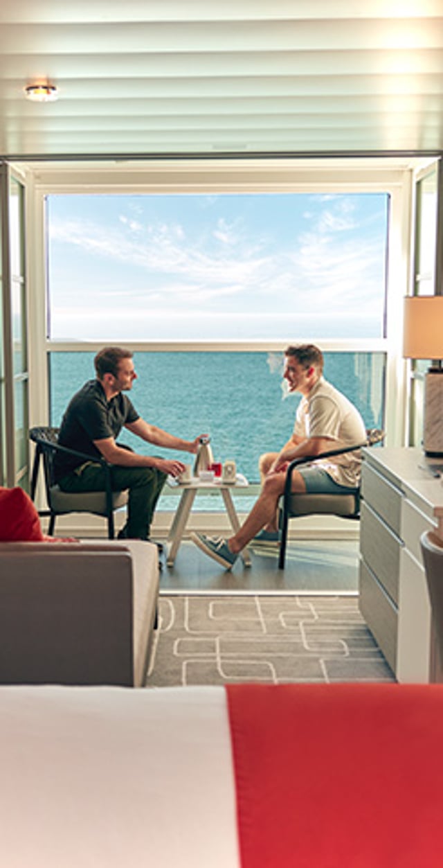 Two men sit and talk in a cruise ship cabin with a large window overlooking the ocean.