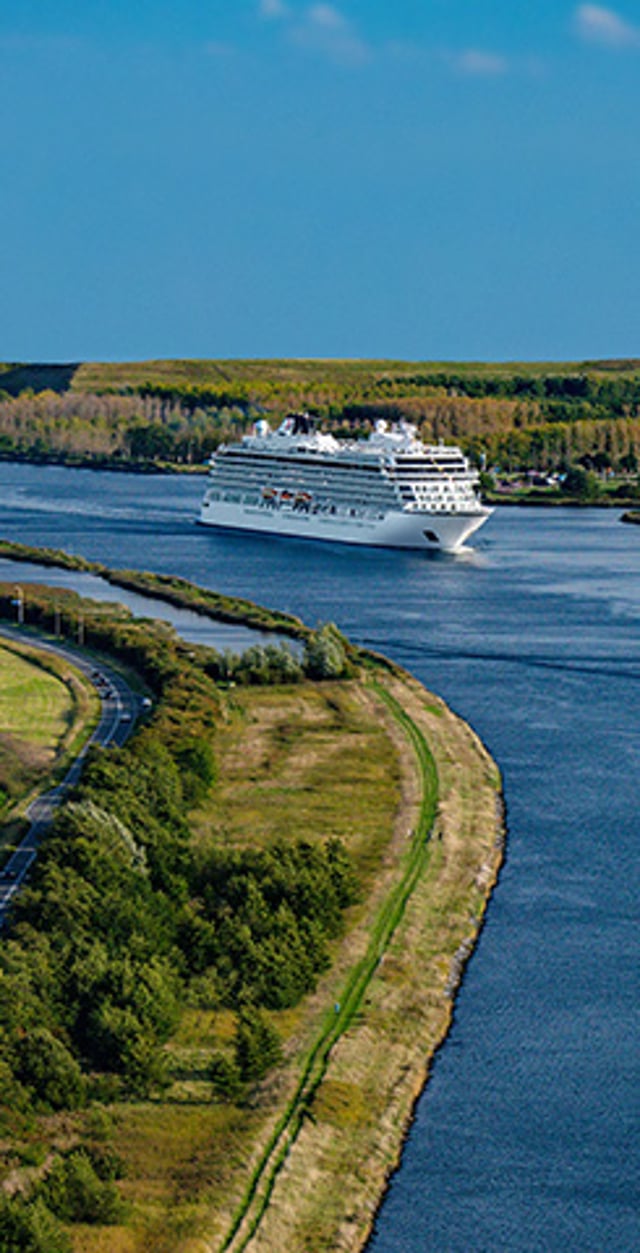 A white cruise ship sails through a narrow river bordered by green fields and forest.