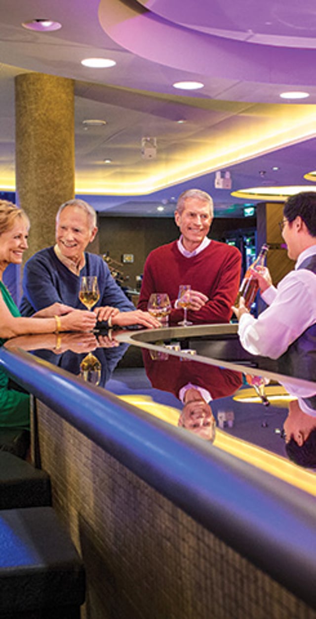 Three cruise guests smiling and talking with a bartender at an onboard lounge.