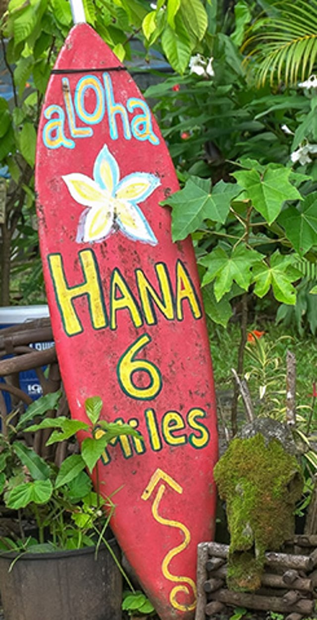 Colorful red surfboard-shaped sign reading 'Aloha Hana 6 miles' surrounded by lush tropical greenery.