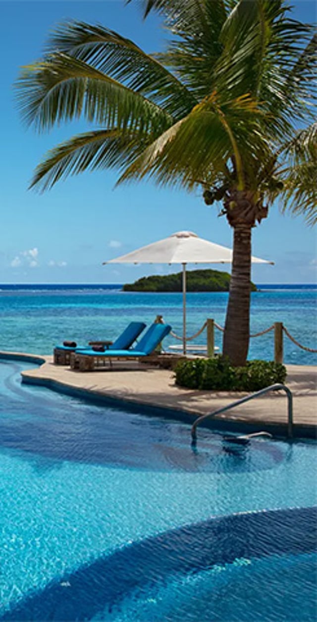 A luxury resort swimming pool next to the ocean, featuring bright blue lounge chairs under a white umbrella, a tall palm tree, and a small island visible on the horizon.