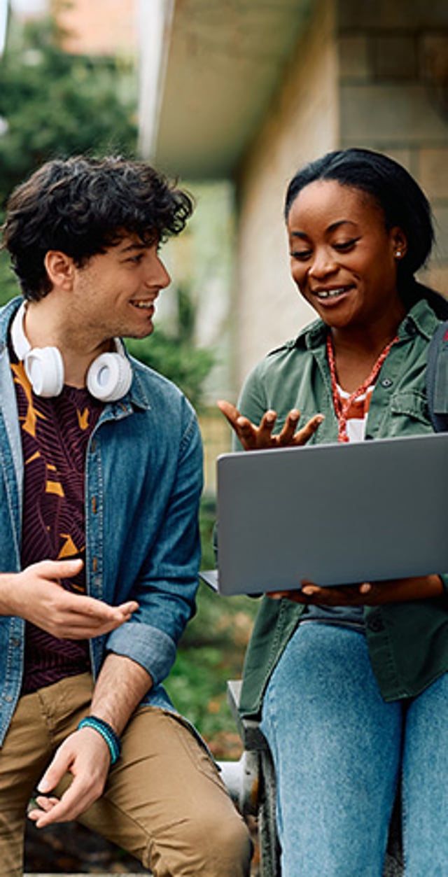 Two college students sitting outside and smiling while looking at a laptop together.