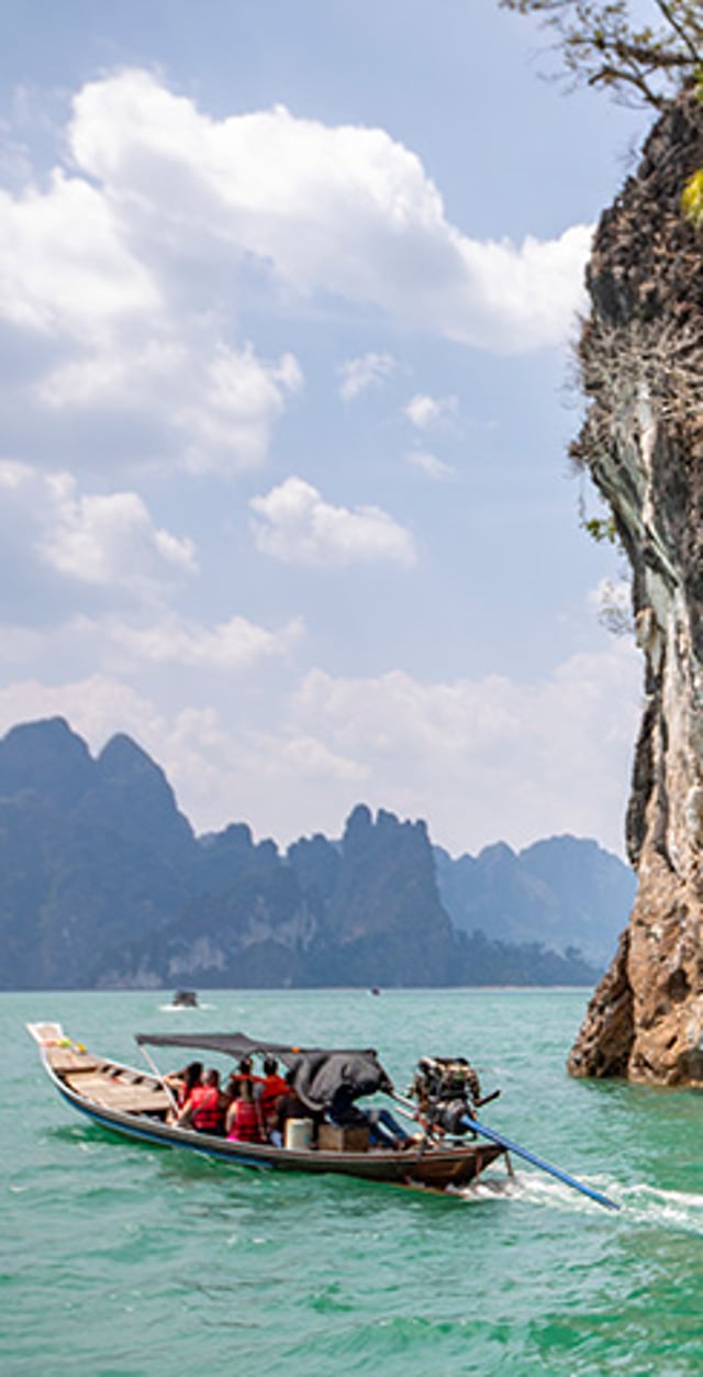 Long-tail boat carrying passengers on turquoise water beside a limestone cliff, with tropical islands in the distance.