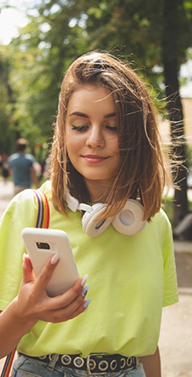 Young woman wearing headphones around her neck and looking at her phone outdoors.