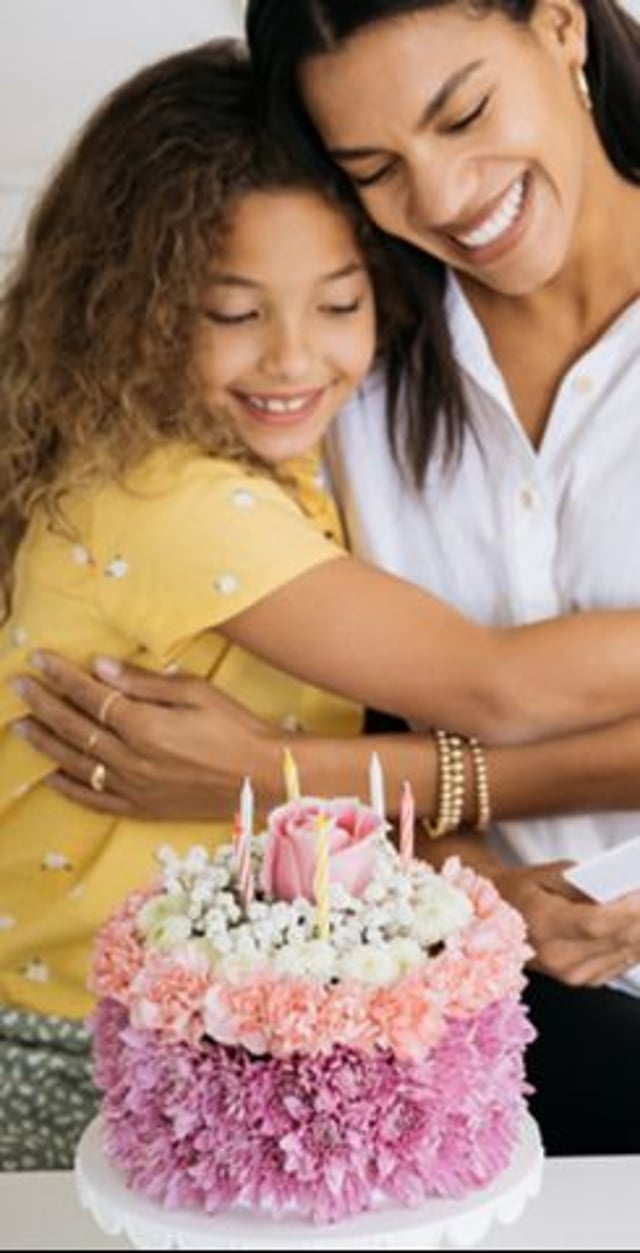 A woman and a young girl embrace, smiling joyfully in front of a decorated birthday cake with candles and pink floral accents.