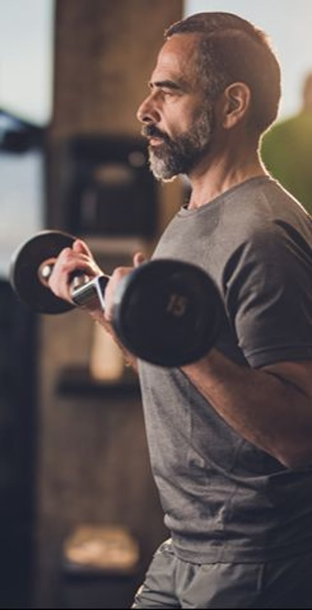 A focused man lifts a dumbbell in a gym setting, dressed in a gray t-shirt and shorts, with natural light illuminating his profile.