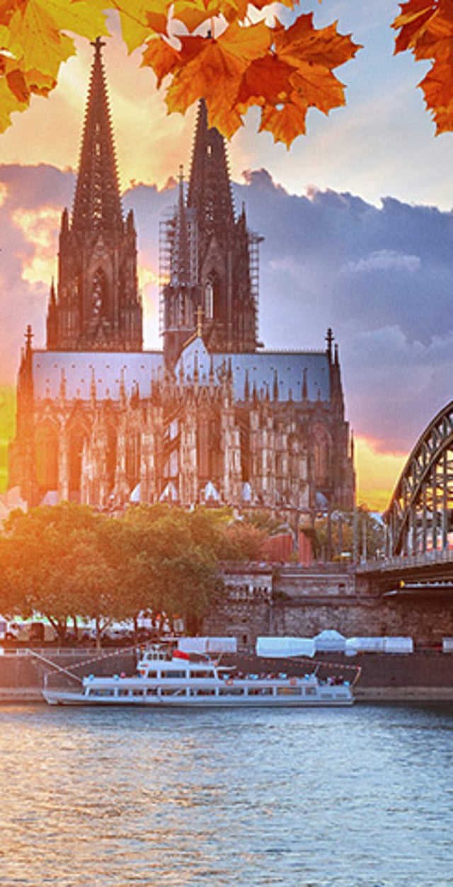 Cologne Cathedral overlooking the Rhine River at sunset, with autumn leaves framing the skyline and a sightseeing boat on the water.