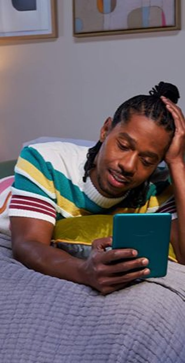 A man in a striped sweater lies on a bed, looking intently at a tablet, with one hand on his head, in a softly lit room.