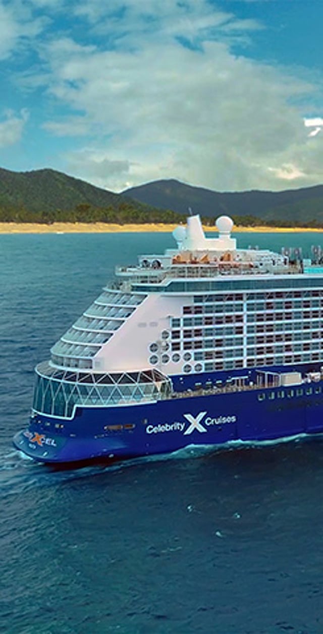 Celebrity Cruises ship sailing on calm blue ocean waters with a tropical island and green mountains in the background.