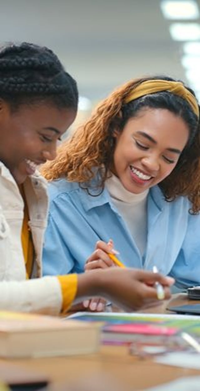 Two young women studying together at a table, smiling. One has short braids and a light jacket, while the other has long hair with a yellow headband. Colorful materials are spread across the table.