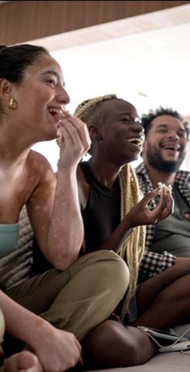 disc-directv-mobileappcoupon-0726.jpg   Image Breakdown Long Description:  The image features three people sitting close together on a floor, engaged in a moment of enjoyment or laughter, suggesting a relaxed social gathering. The setting appears to be indoors, with soft, natural lighting illuminating their faces. The first person on the left has long, dark hair and wears a light blue top, with visible skin texture variations, indicating signs of vitiligo. The second person, in the center, has medium-length blonde braided hair and is wearing a sleeveless black top. The third person, on the right, has a beard and a cheerful expression, dressed in a green shirt with a checkered pattern visible in the background. They each are holding snacks, suggesting they are sharing a casual moment together.  Alt-text:  Three young individuals laughing and sharing snacks while sitting on the couch indoors, showcasing a relaxed social moment.