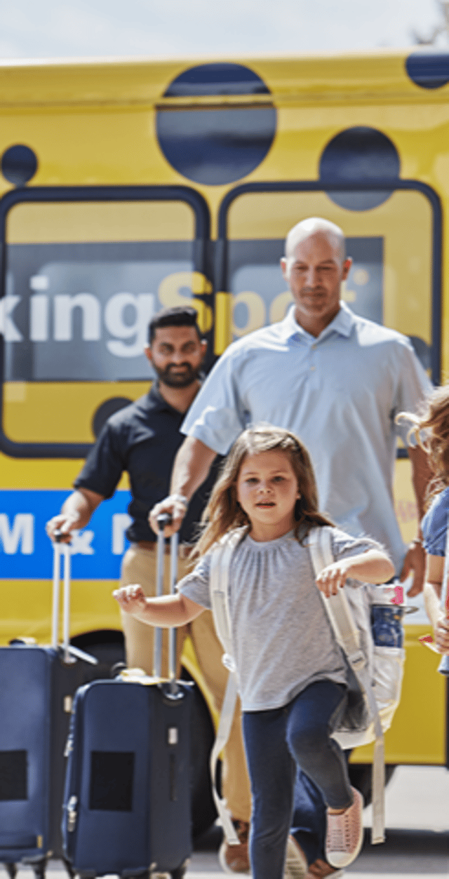 A family descends from a yellow shuttle, with a girl running ahead excitedly, while two men carry luggage.