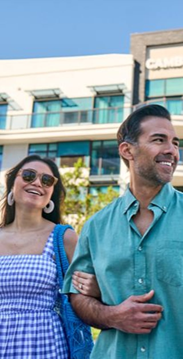 A smiling couple strolls together outdoors; the woman in a gingham dress and sunglasses, and the man in a turquoise shirt, with a modern hotel in the background.
