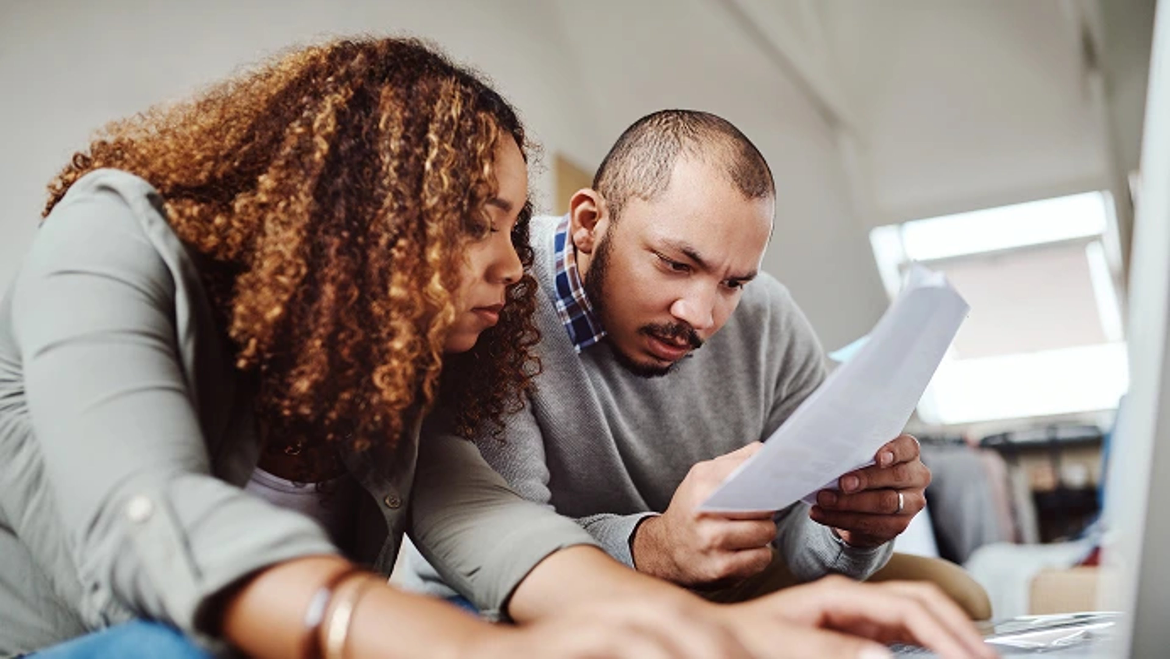 A man and a woman with serious expressions leaning in closely to review a paper document together while working on a laptop