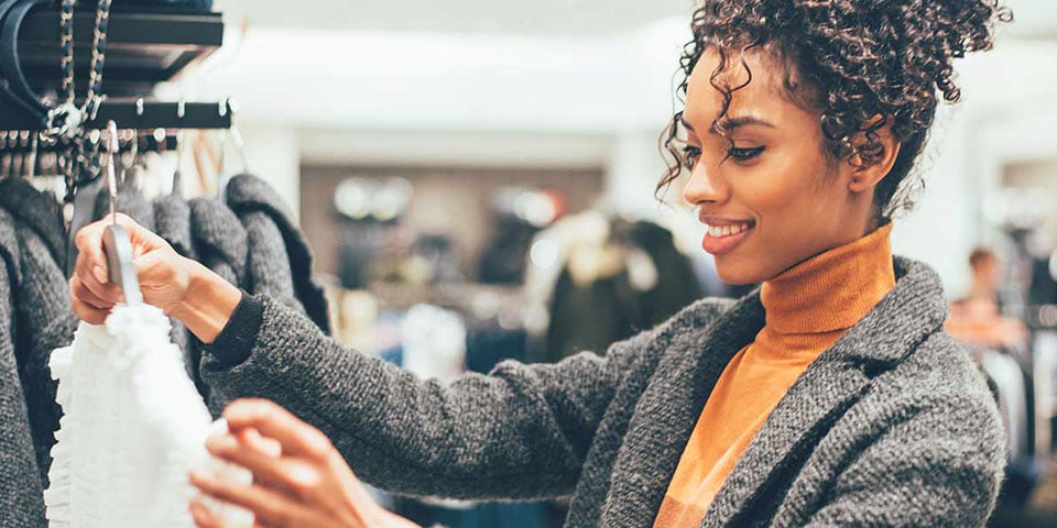AAA Discounts Shopping and Retail Young woman smiling as she holds up white garment on hanger while shopping in clothing store