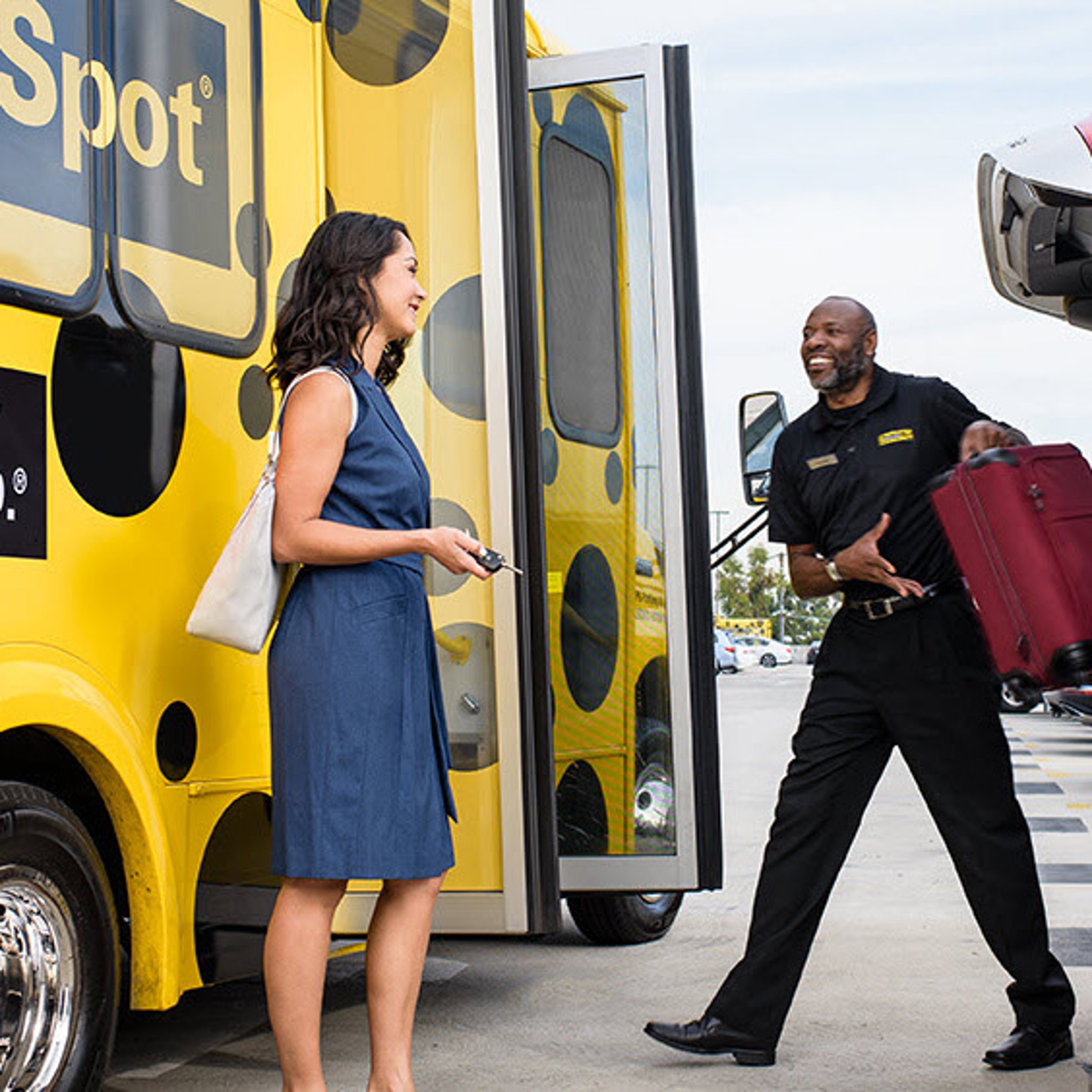 Driver for The Parking Spot smiling as he carries a woman's red suitcase as she is about to board yellow shuttle with black polka dots