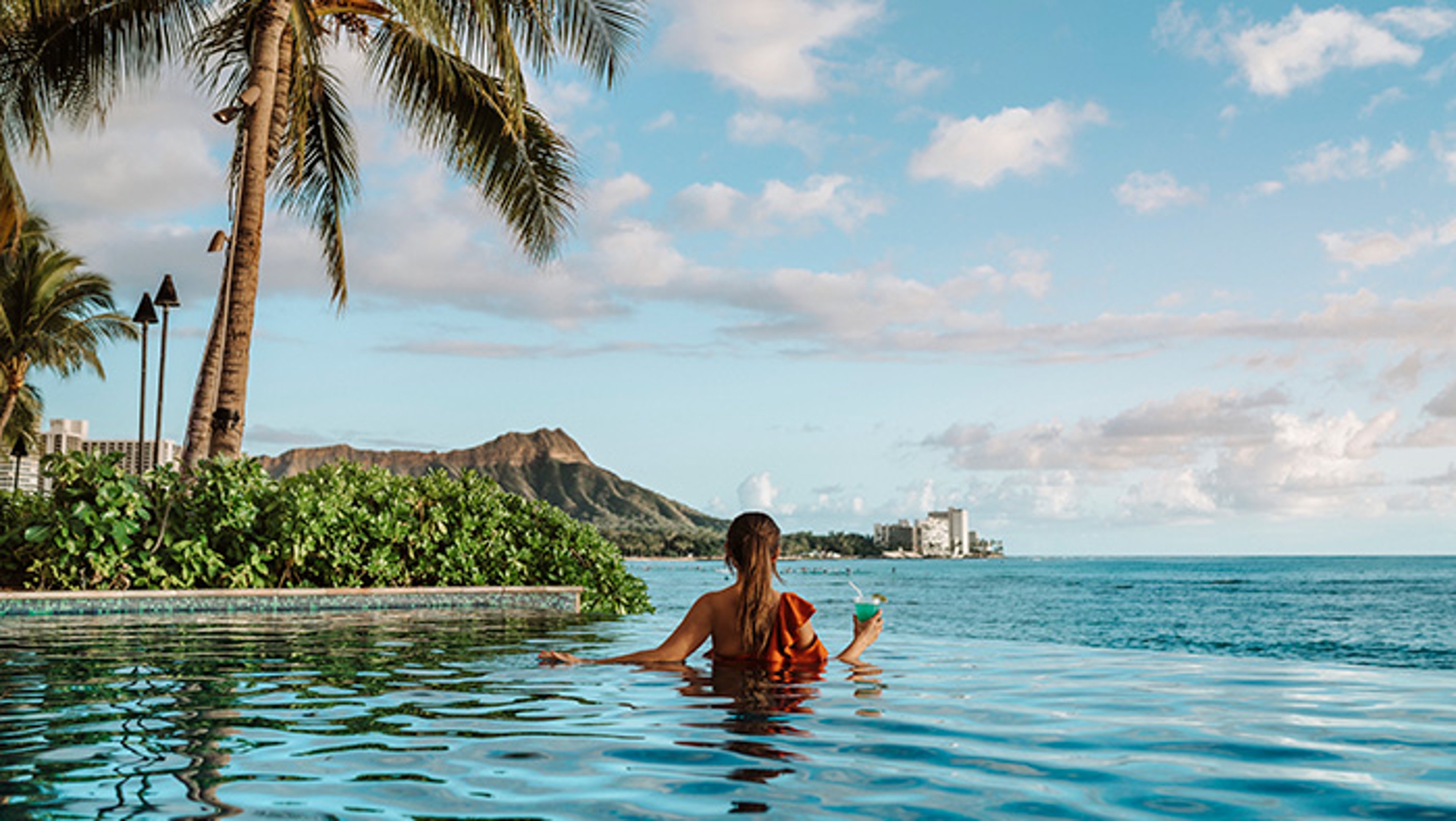 Person in an infinity pool overlooking the ocean and a mountain, with palm trees nearby.