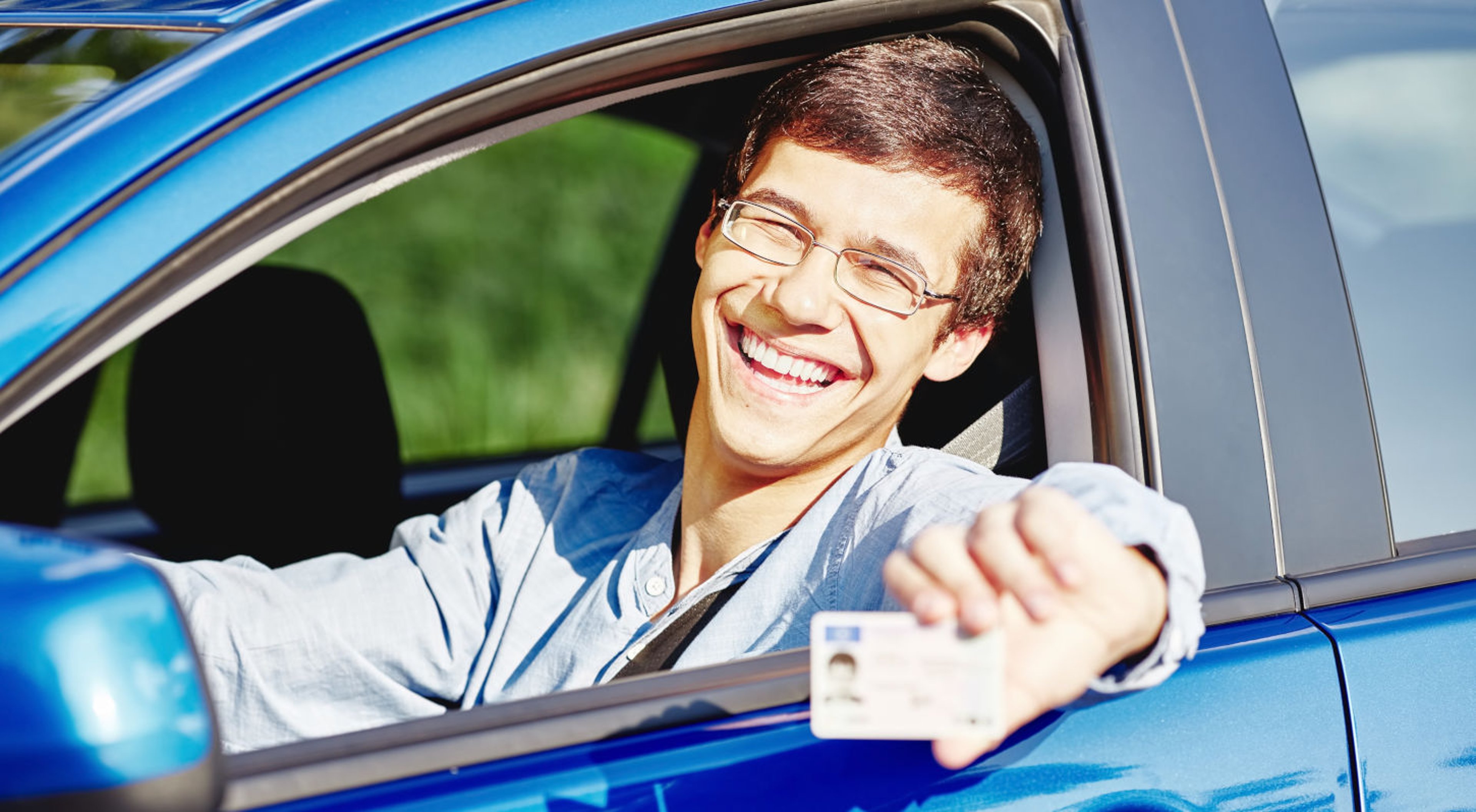 Teenage boy driver in blue car smiling in driver's seat as he shows his license through open window