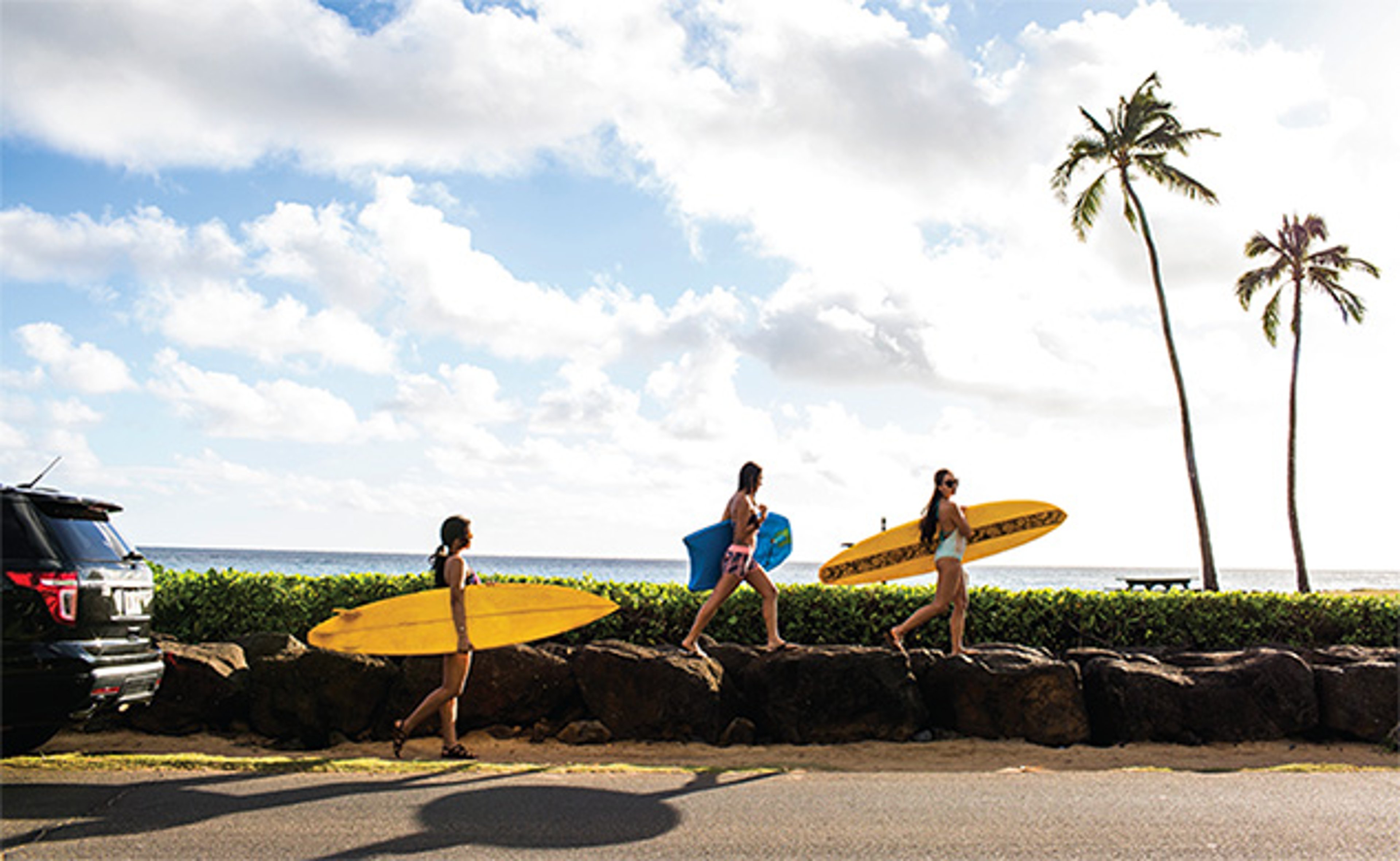 Family walking with surfboards along a coastal road next to an SUV, with ocean and palm trees in the background.
