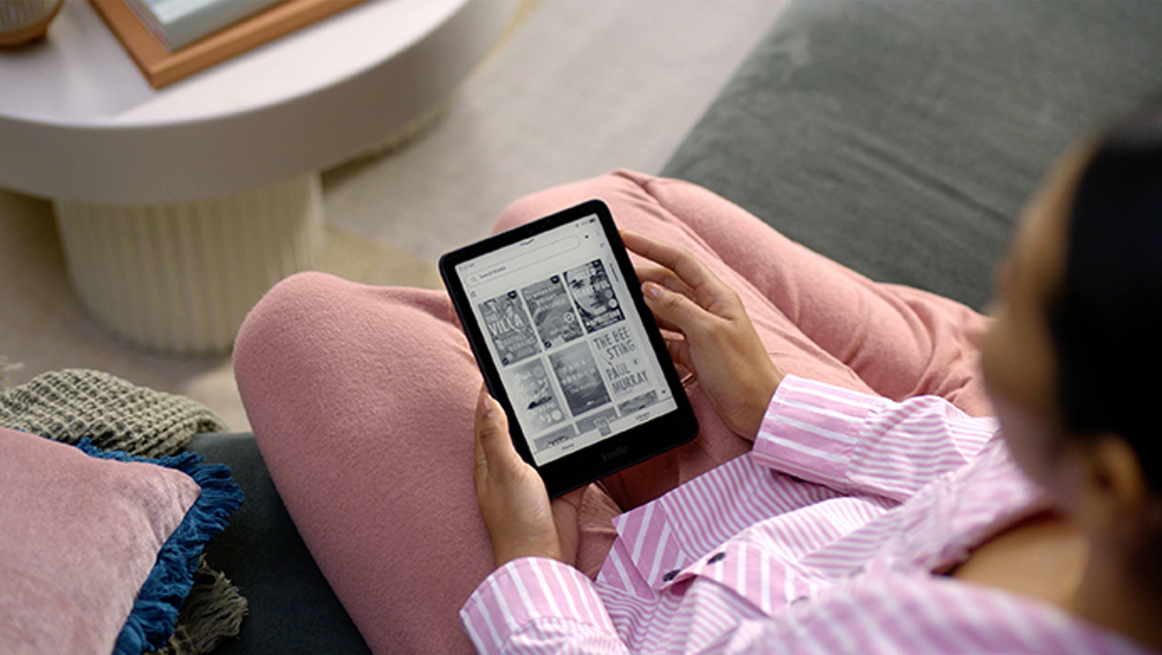 A person in pink pajamas is reading on a tablet while sitting on a couch, with a coffee table and framed picture nearby.