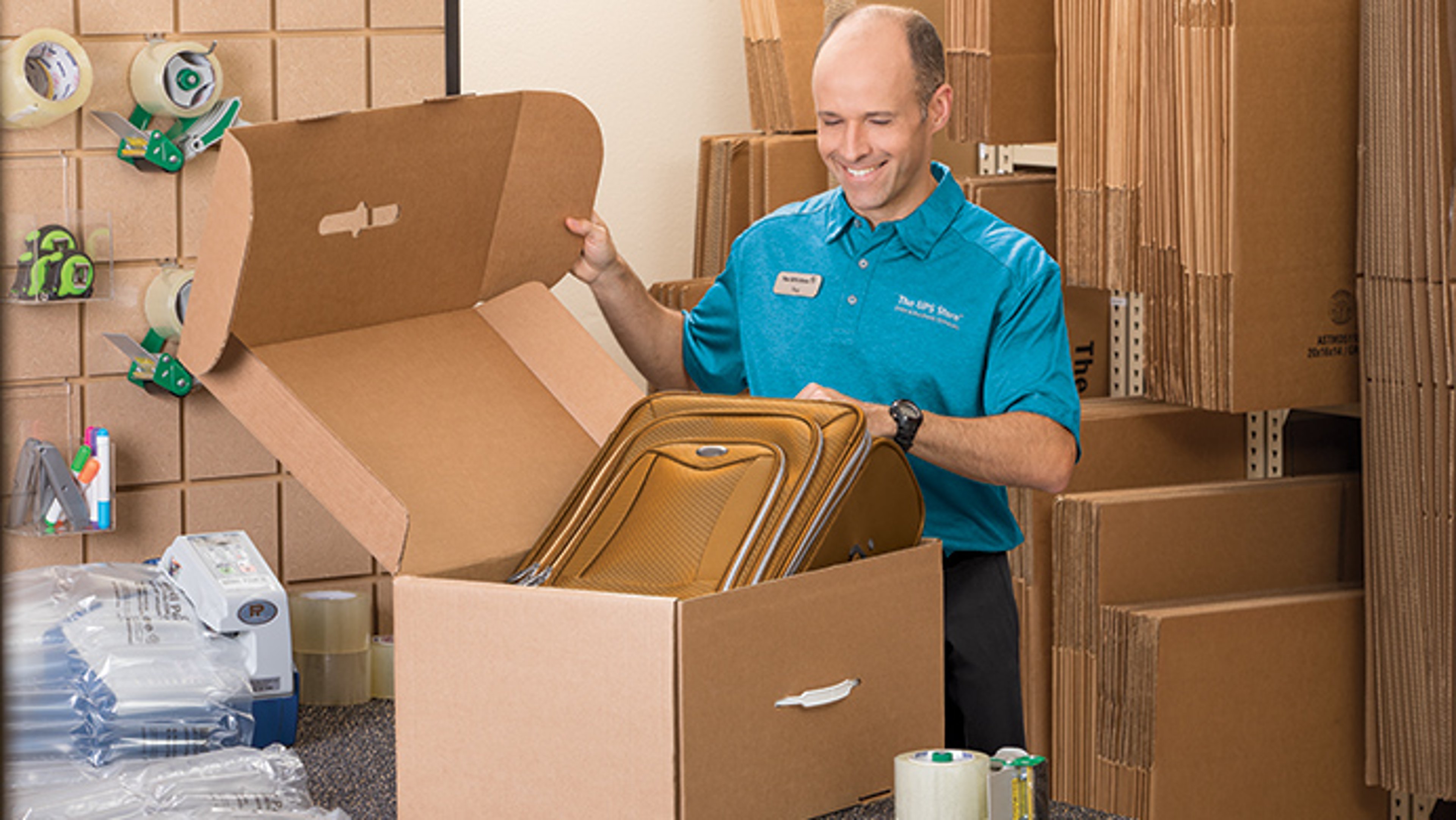 A man in a blue shirt smiles as he places a brown suitcase into a large cardboard box, surrounded by packing materials and cardboard boxes.