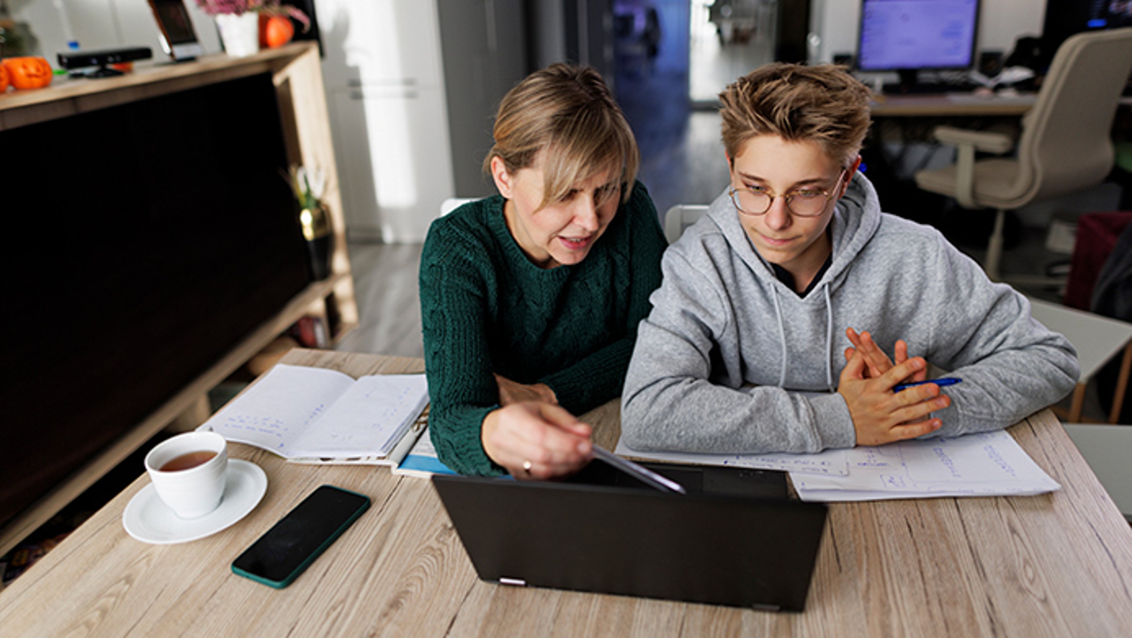 A woman in a green sweater and a young man in a grey hoodie engage with a laptop in a cozy study space, with books and a cup of coffee on the table.