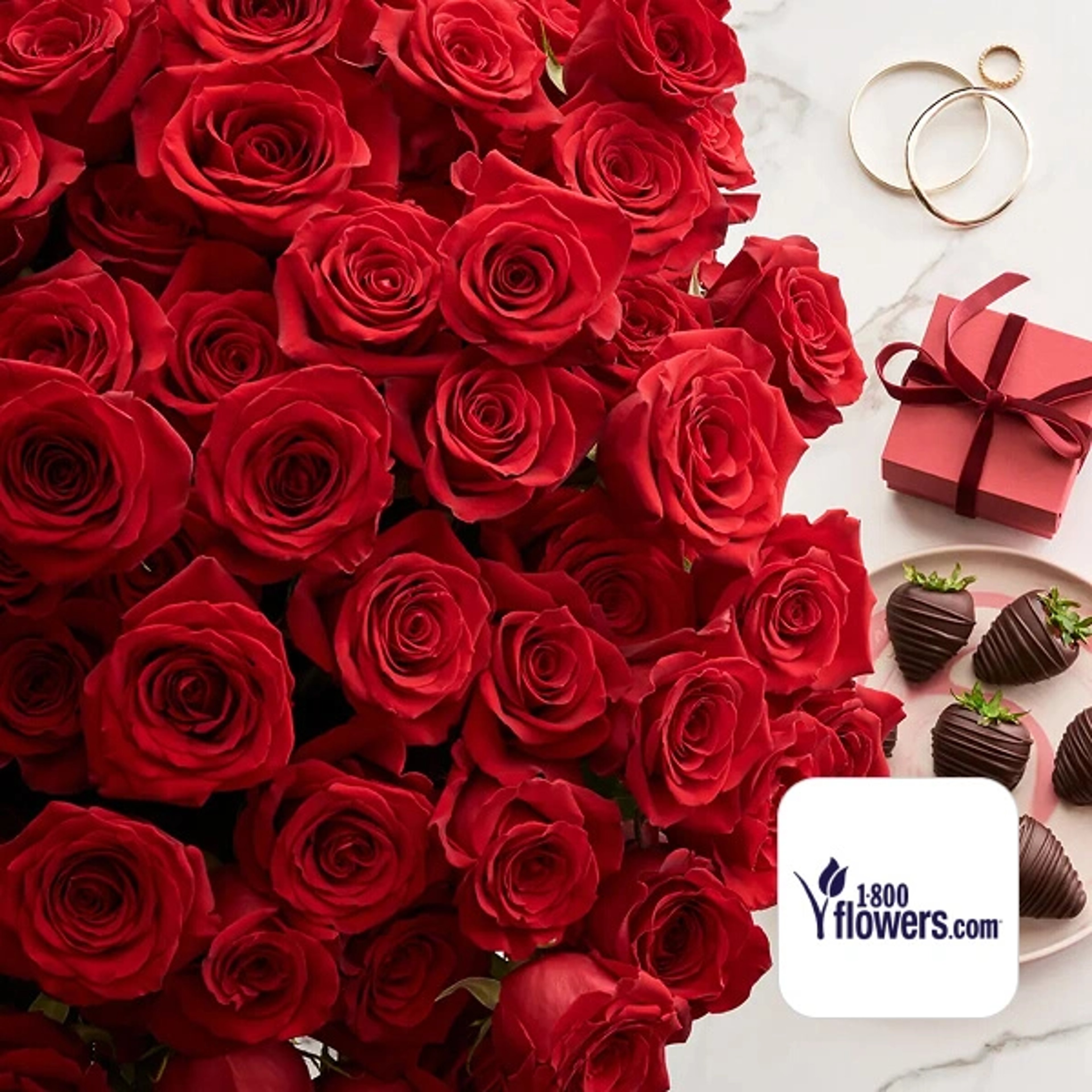 A large, lush bouquet of vibrant red roses next to a red gift box with a ribbon, a few gold rings, and a platter of chocolate-covered strawberries, with the 1800flowers.com logo in the corner.