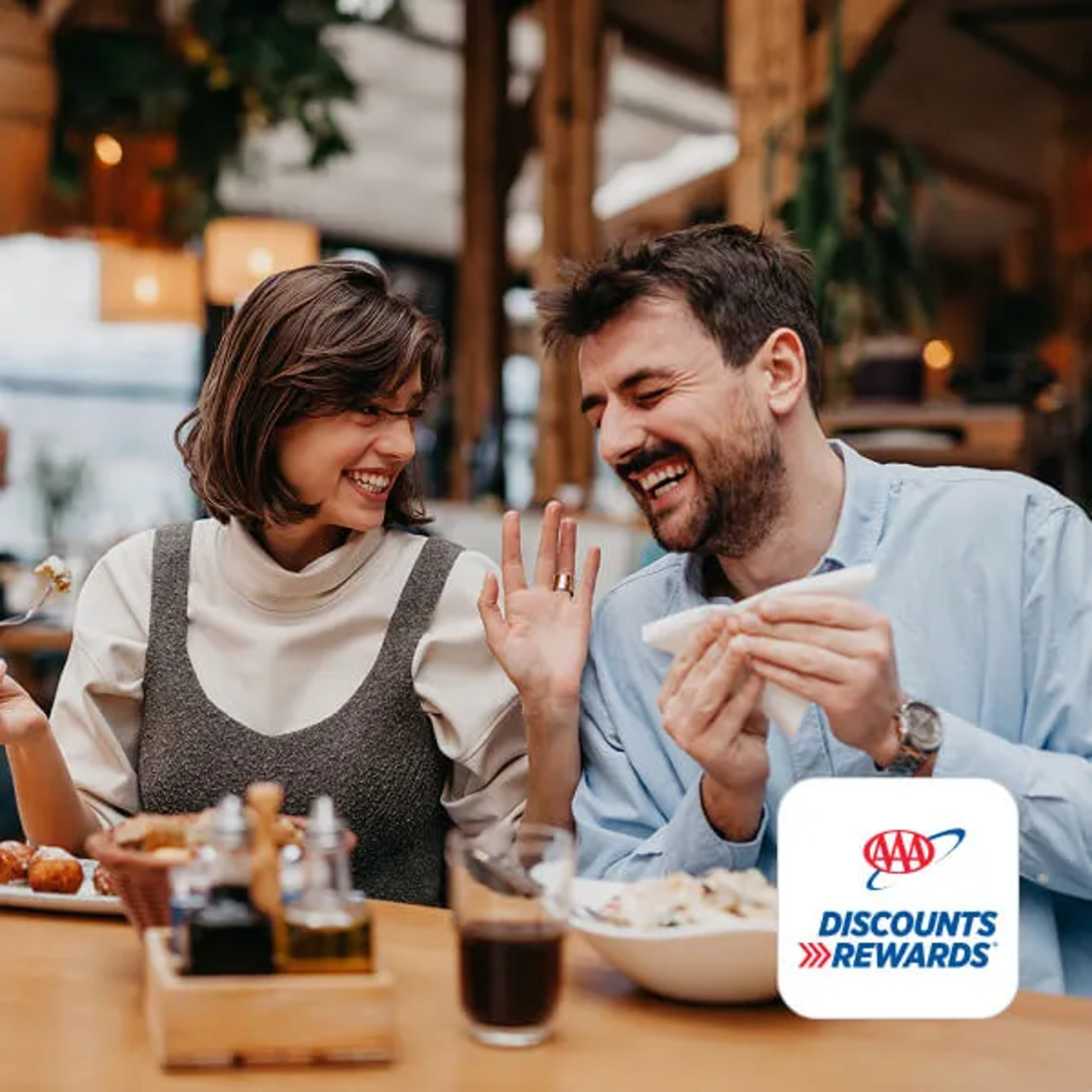 A man and a woman smiling and laughing together over a meal at a restaurant, with the AAA Discounts and Rewards logo in the bottom corner.