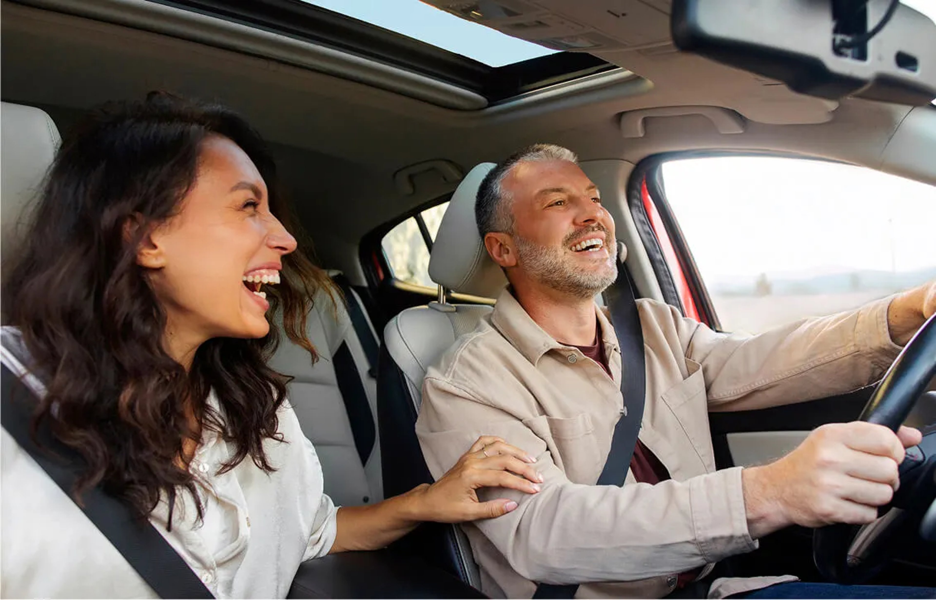 A happy couple laughing together inside a car with an open sunroof during a sunny road trip.
