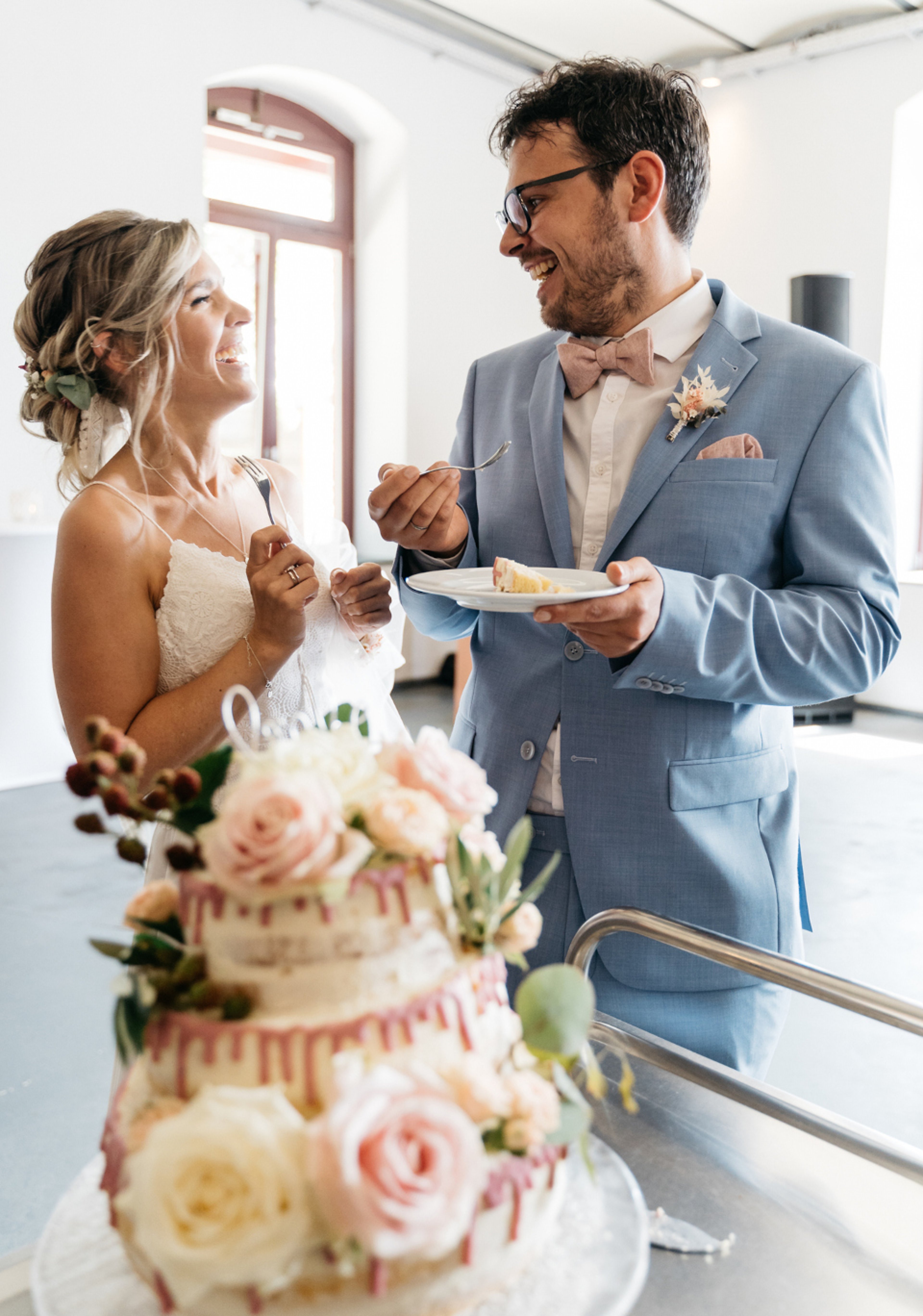 Bride and groom smiling at each other in front of wedding cake decorated with flowers