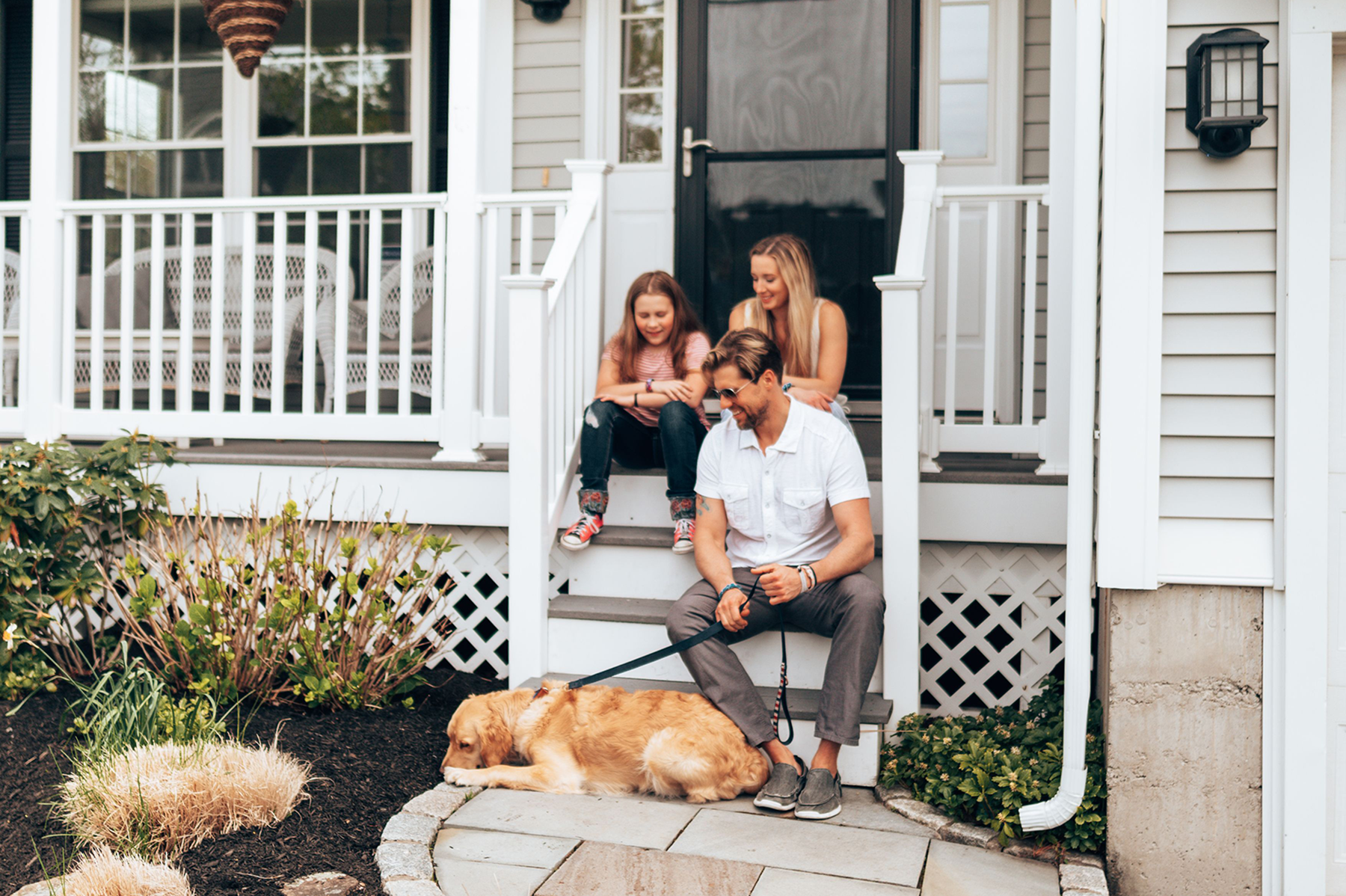 Family of three-mother, father and daughter, sitting on the stoop in front of their house with their dog laying on the ground contently