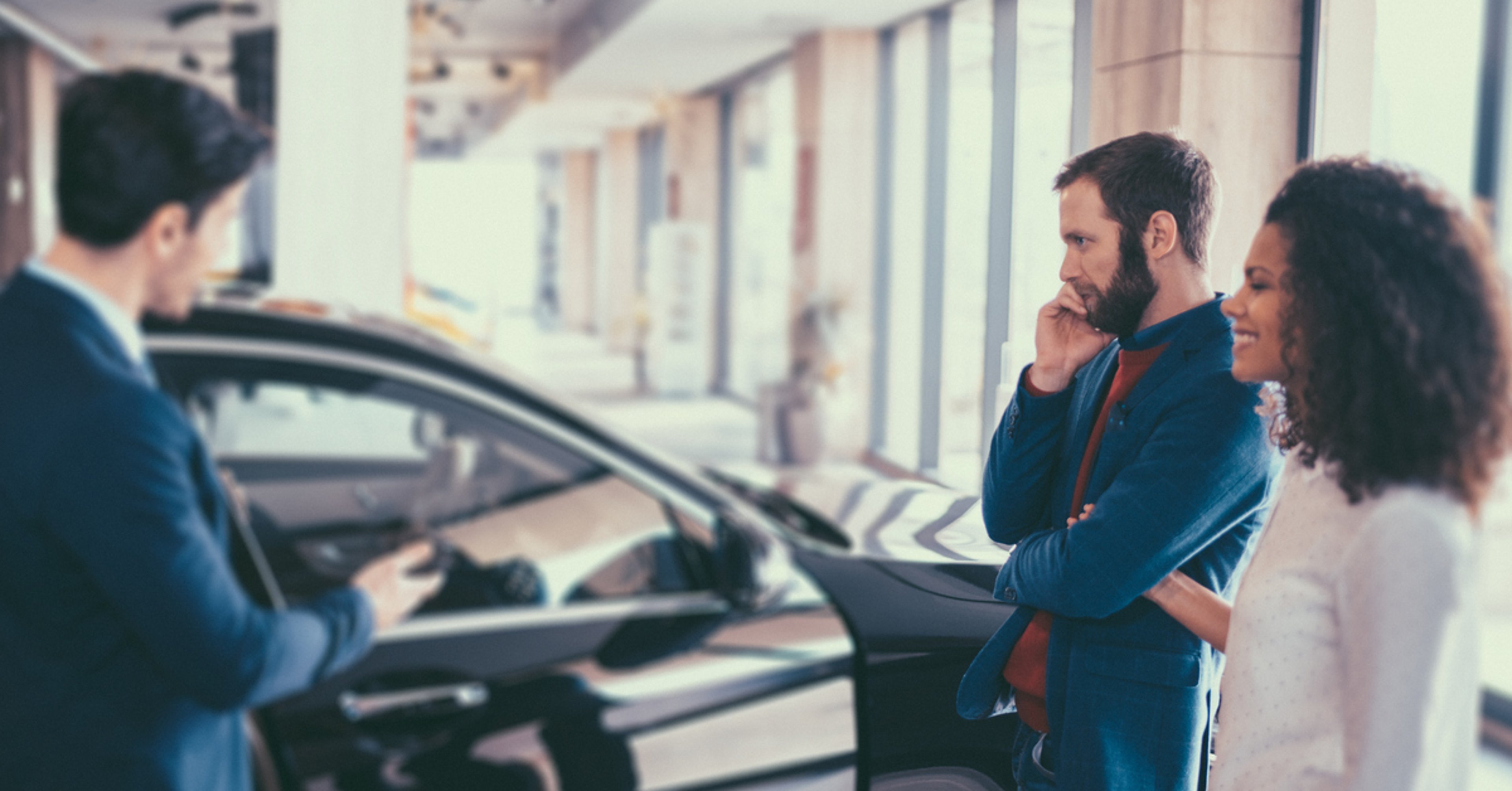 Couple in showroom talking to car salesperson about purchasing a new vehicle with black sedan in background