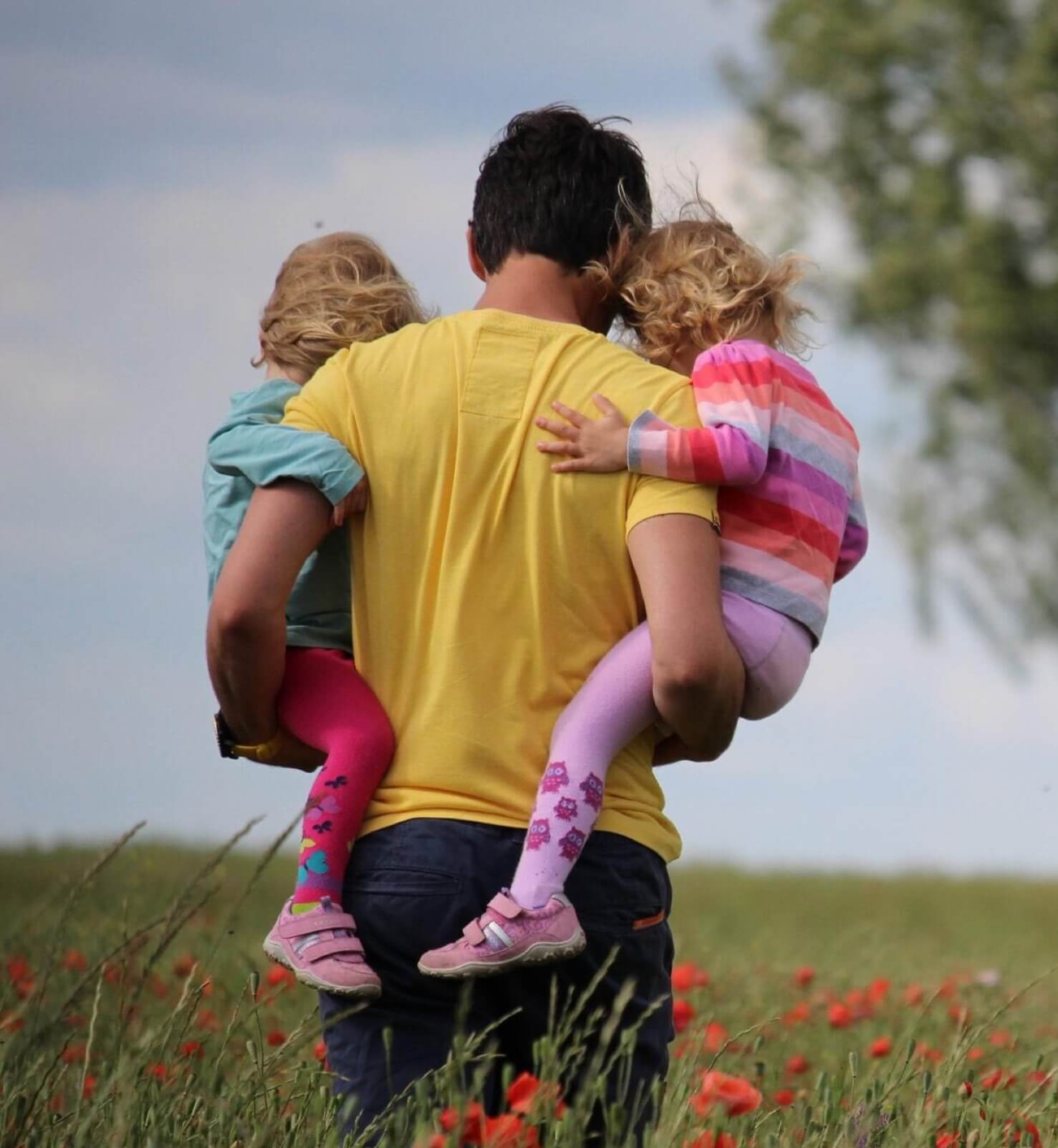 Man with yellow shirt holding two young daughters while standing in field