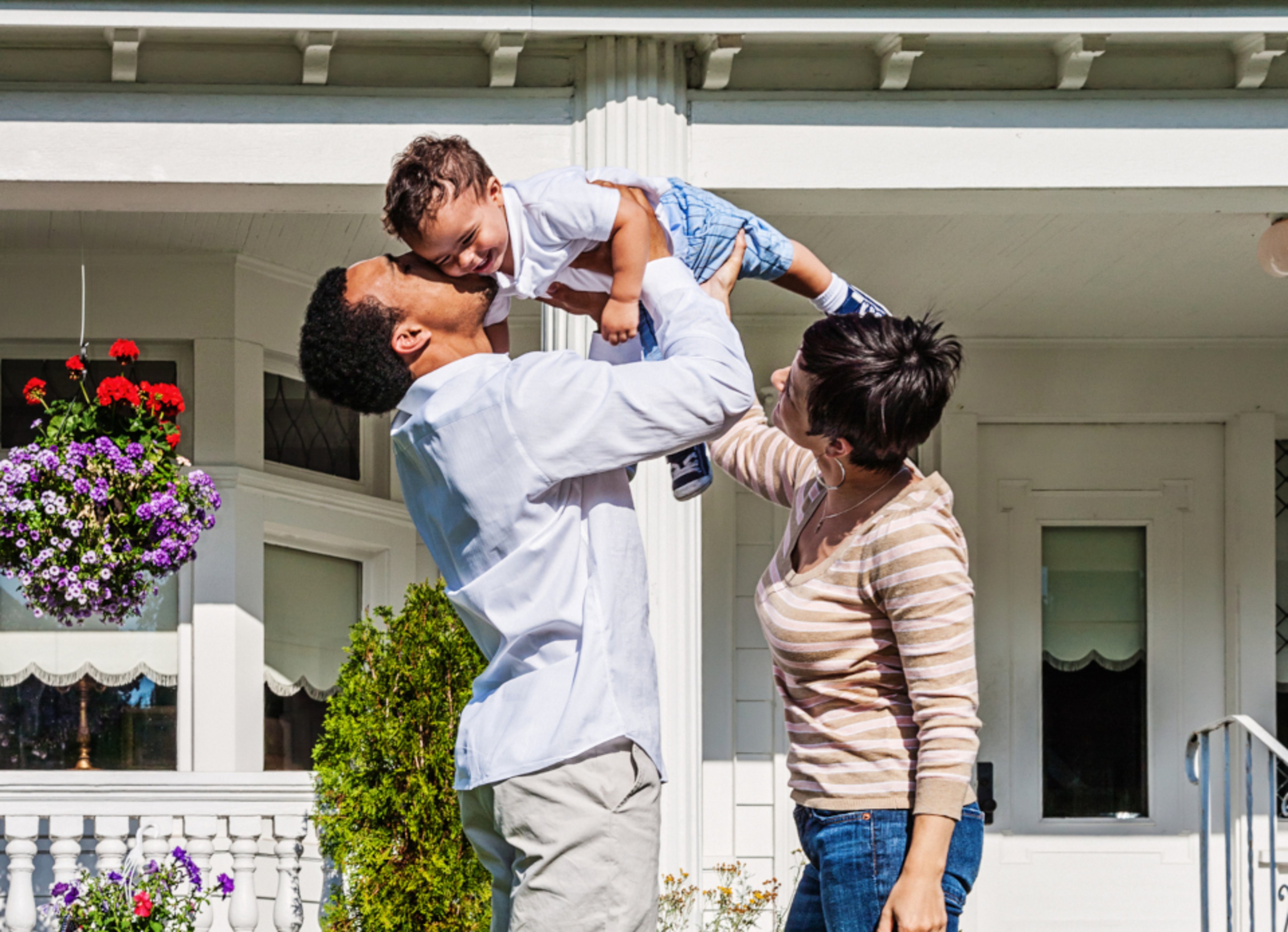 Father holding small child in air outside of home while mother looks at them happily