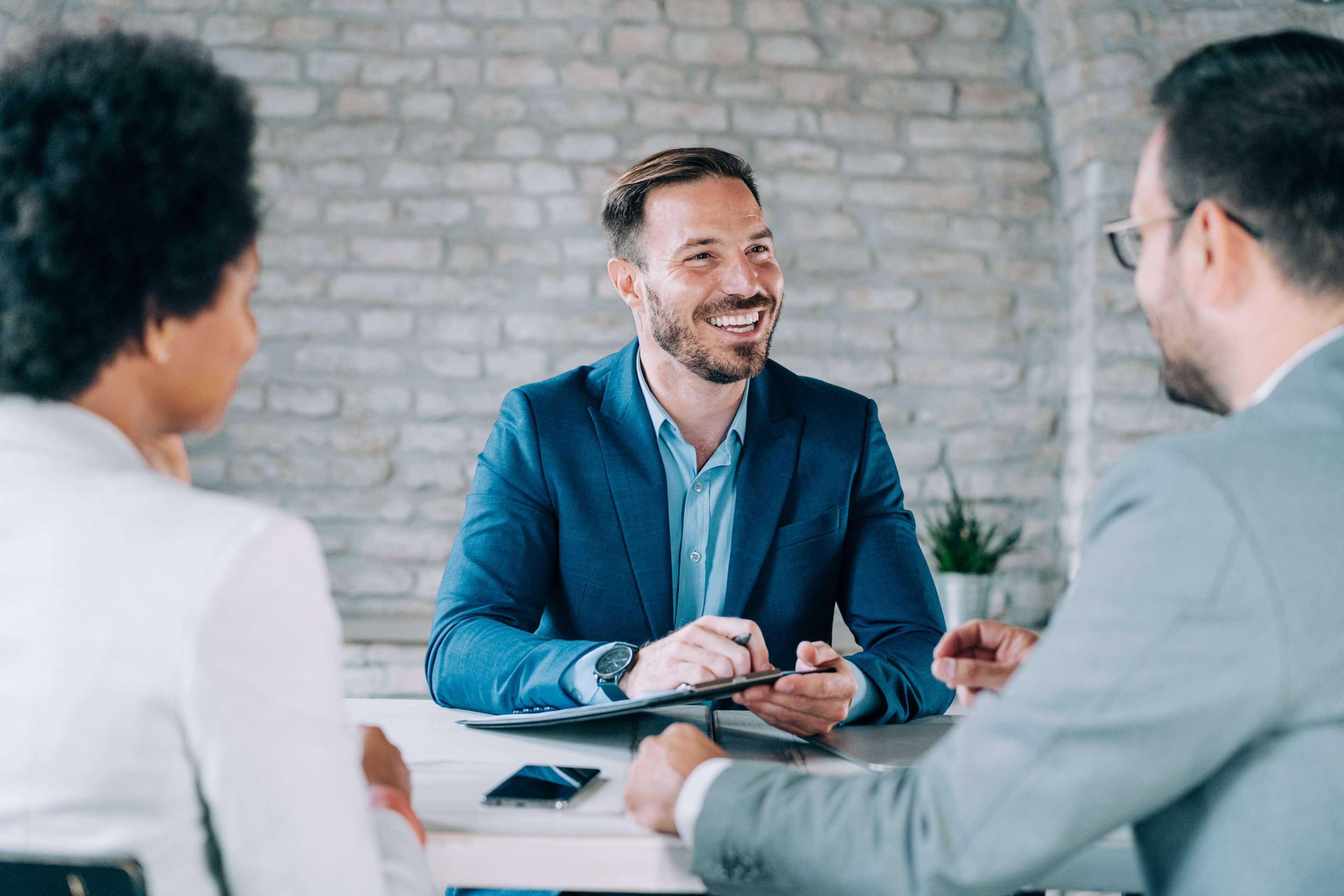 Smiling insurance agent in a blue suit discussing services with two clients in a professional meeting setting.