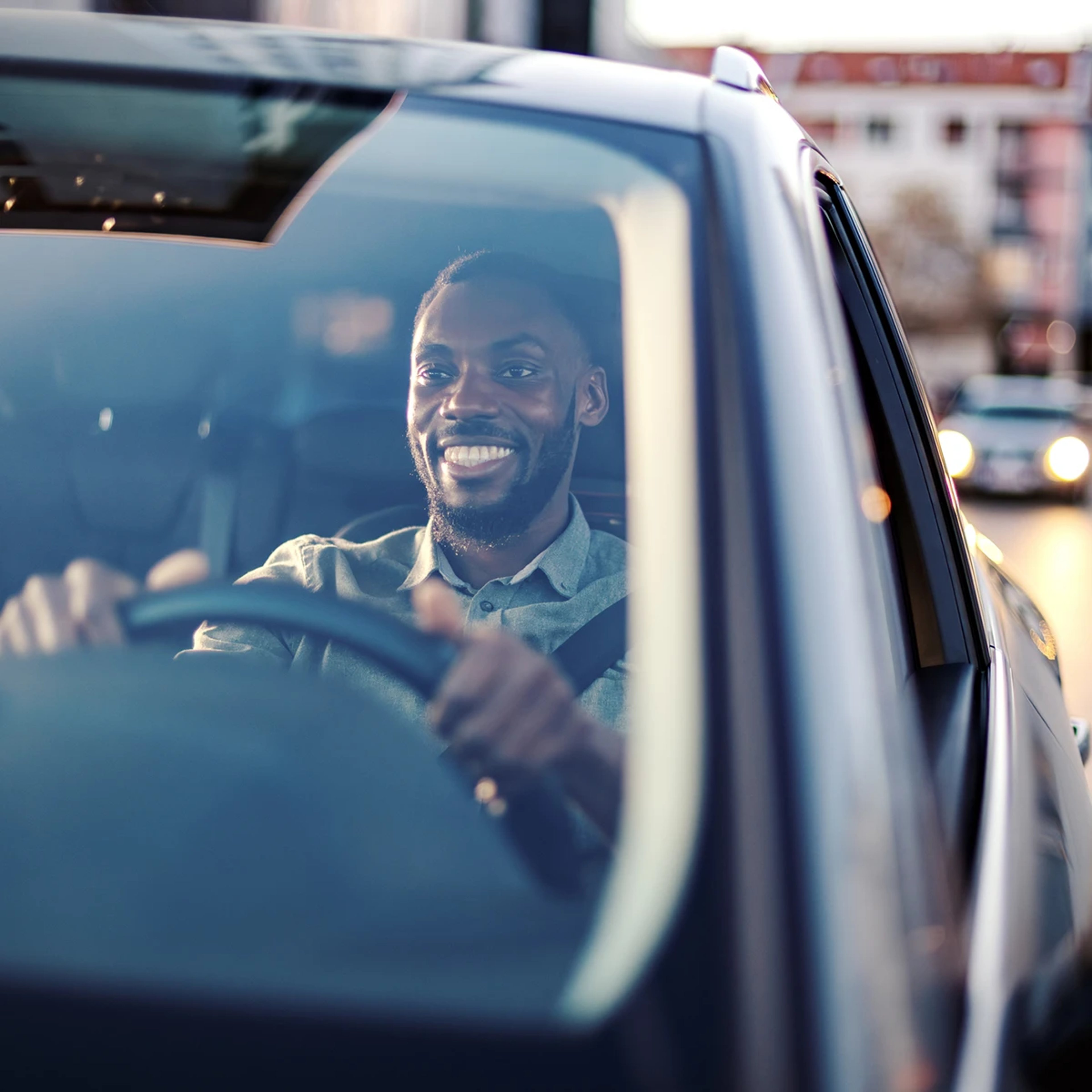 Photo taken of smiling man driving car with cityscape reflecting on windshield