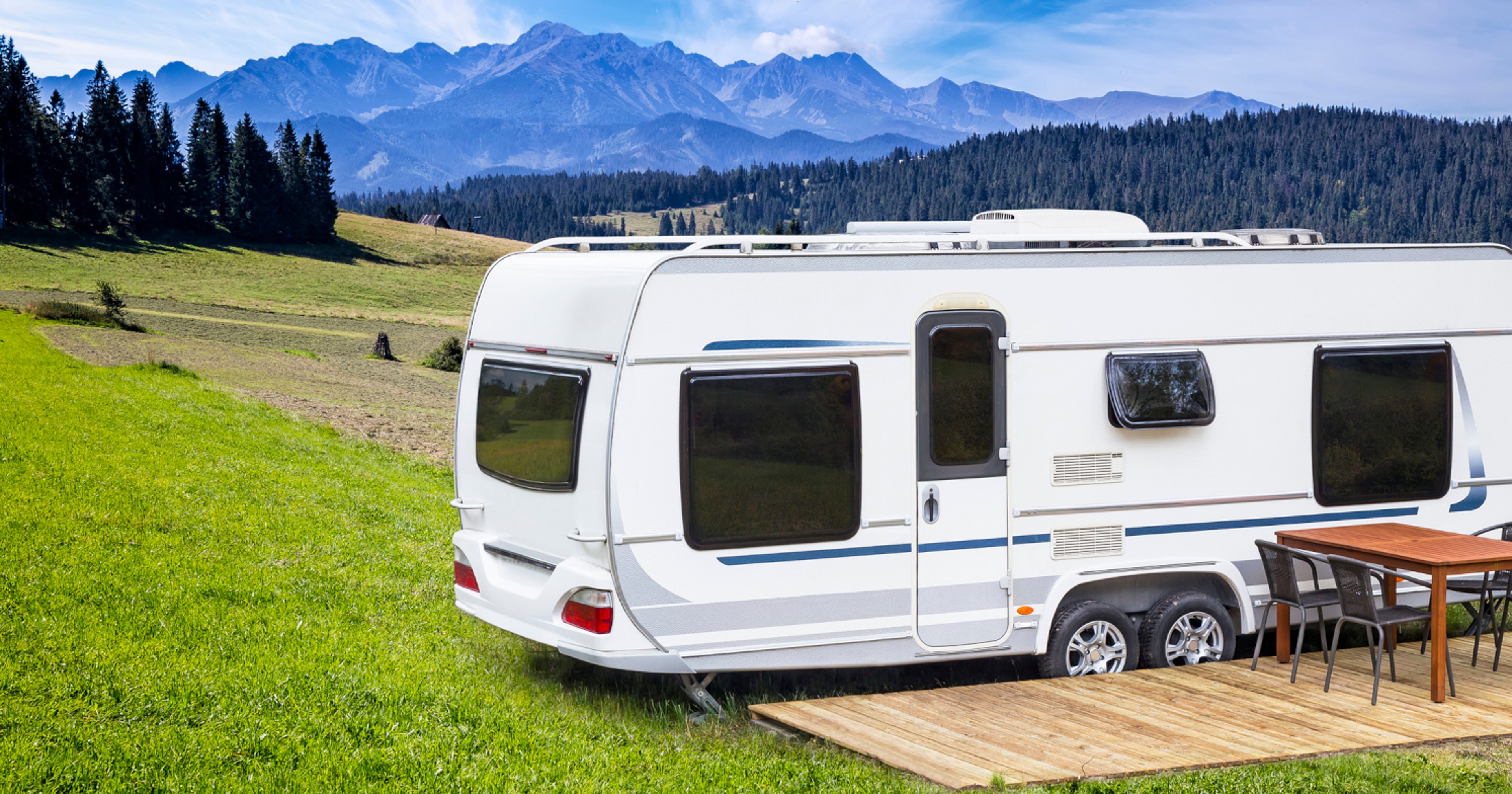 RV parked next to wooden platform with table on lush green hill, surrounded by mountains and trees 