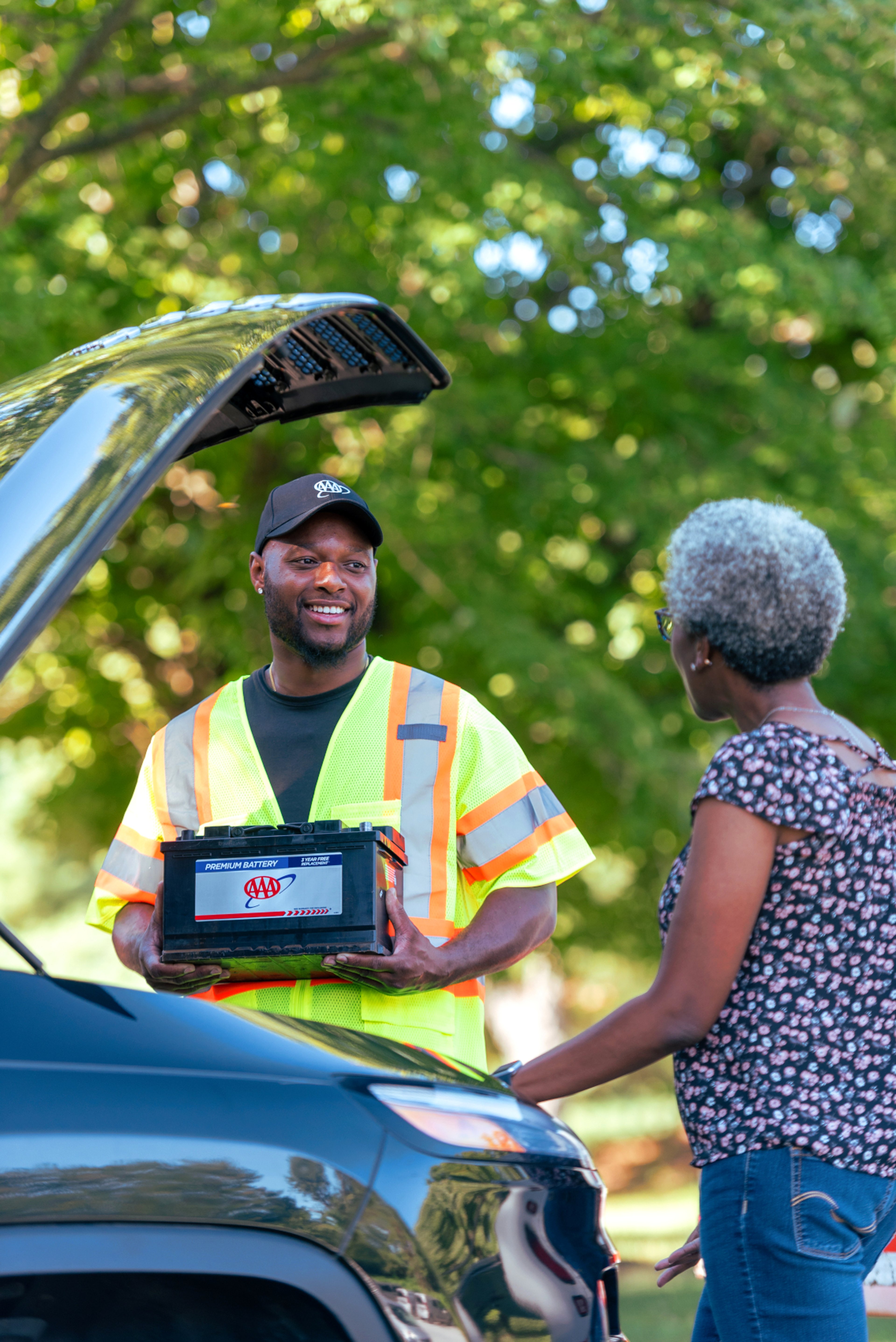 Friendly AAA roadside technican replacing battery standing by open car hood talking to AAA member