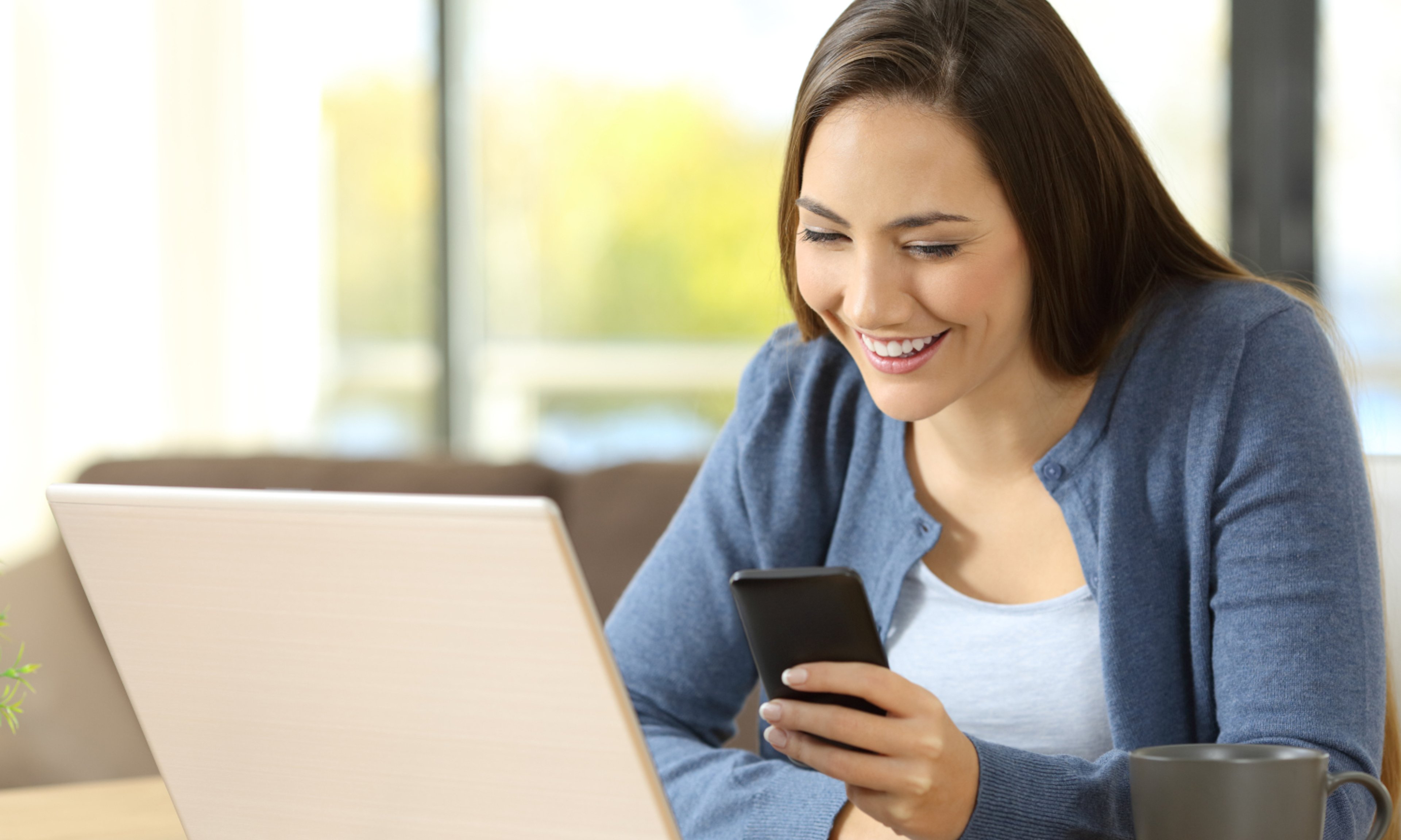 AAA Digital Membership Hero Woman in blue sweater looking at smartphone while browsing on laptop