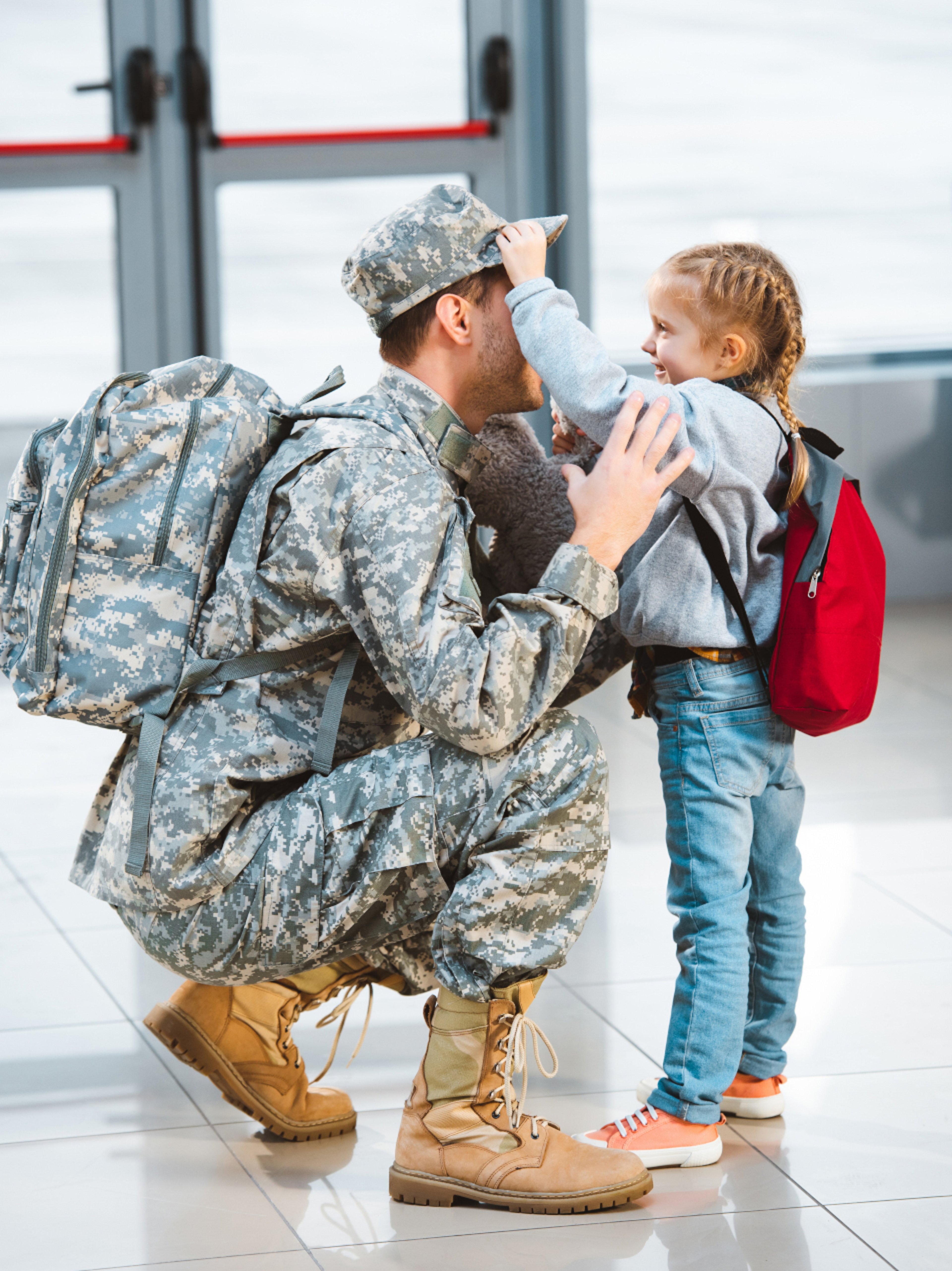 Young daughter with red backpack touching cap of father wearing military uniform