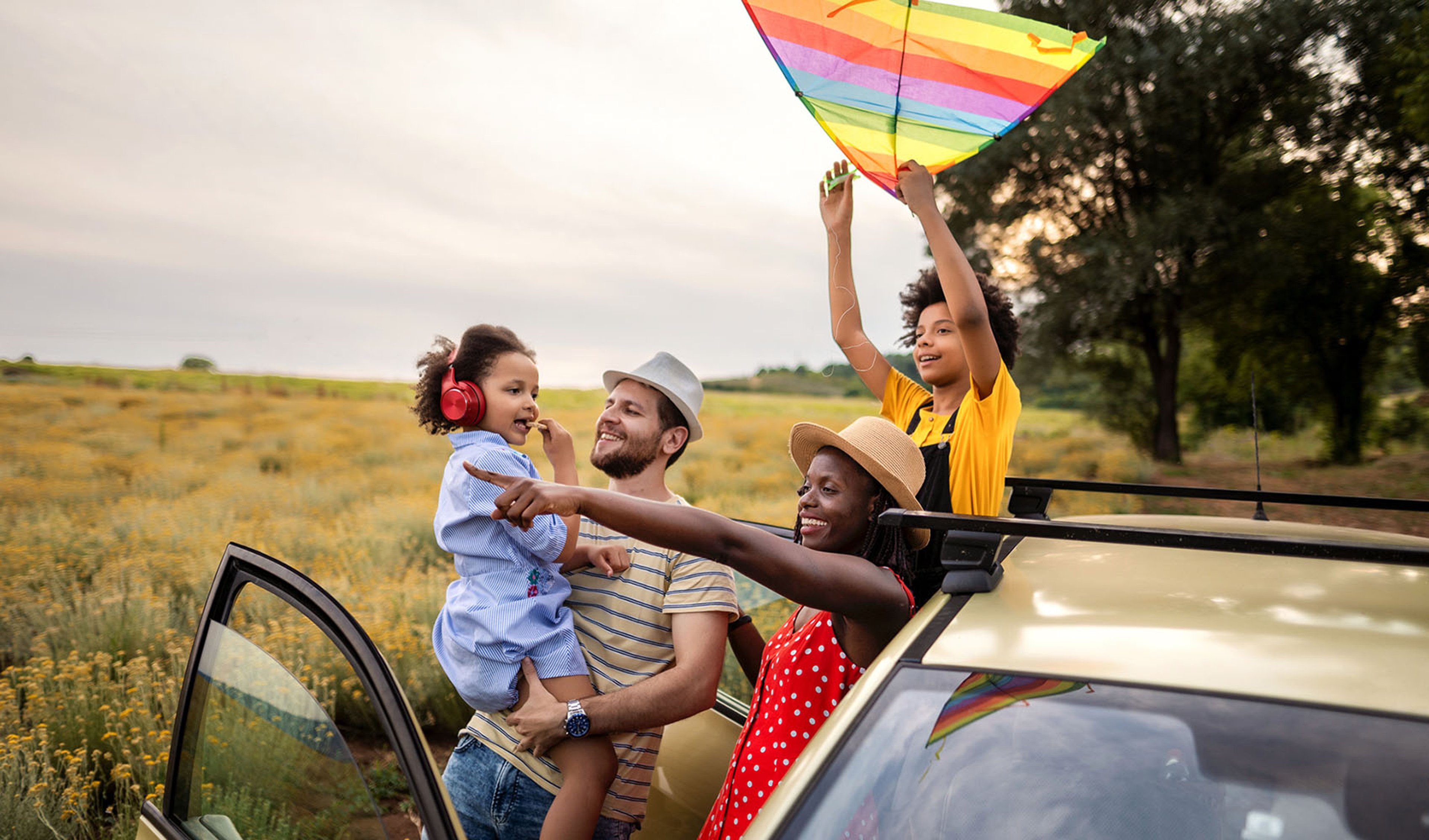 Family of four on vacation standing outside of their vehicle in sprawling field, with child holding a rainbow-colored kite