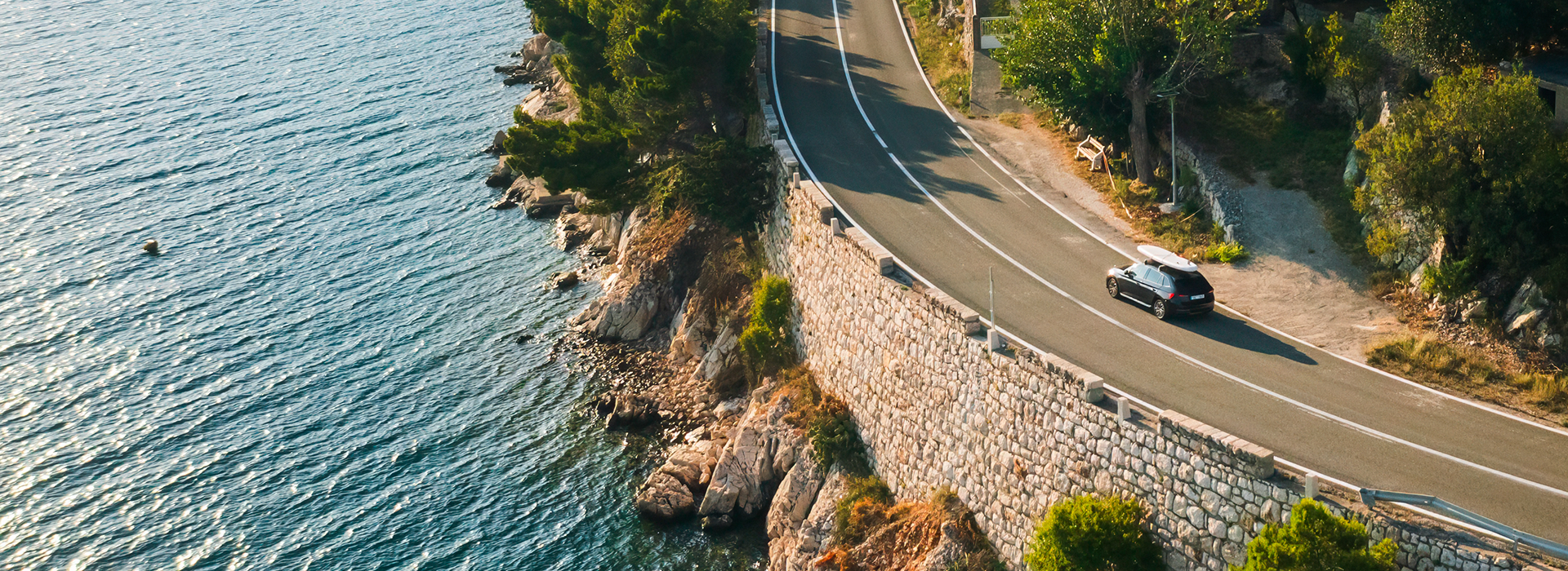 Car with paddleboard on the roof drives along a narrow coastal road under a clear blue sky
