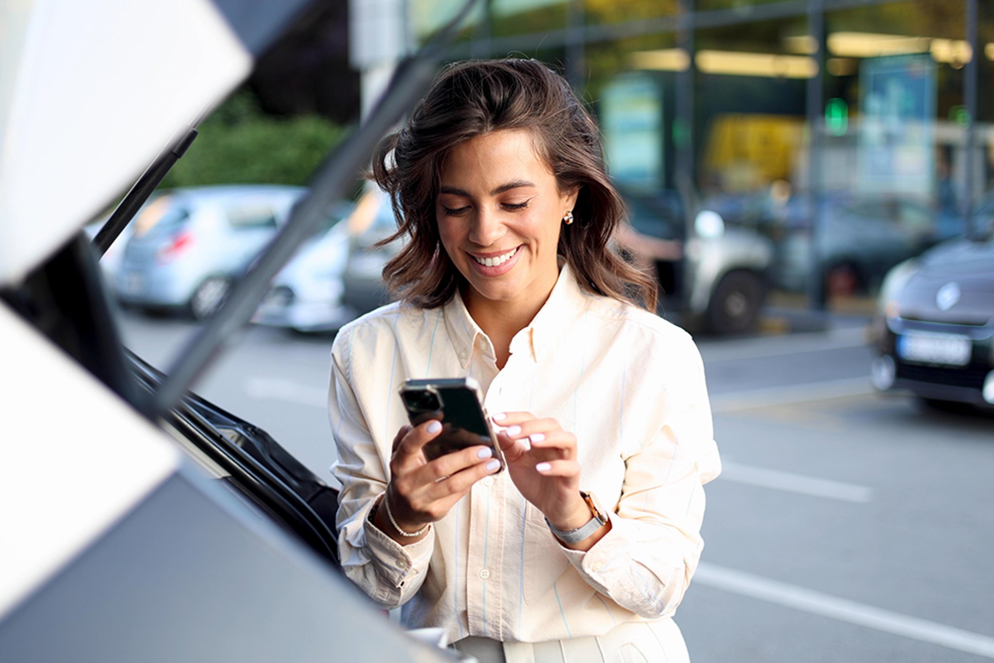 Smiling woman looking down at her phone while standing by her car in a parking lot