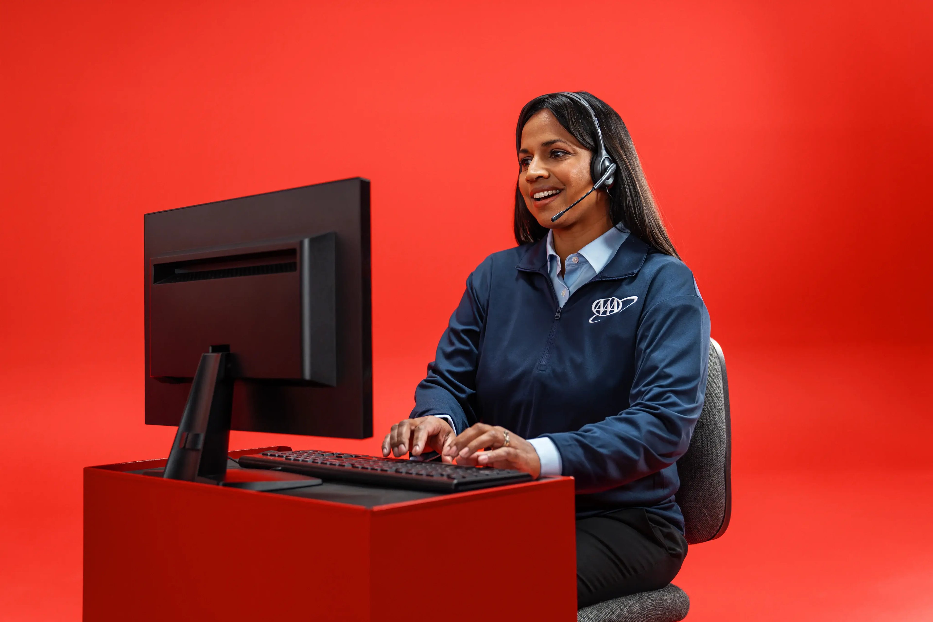 A customer support representative wearing a headset and a dark navy AAA pullover, typing on a keyboard at a computer desk while smiling against a solid red backdrop.