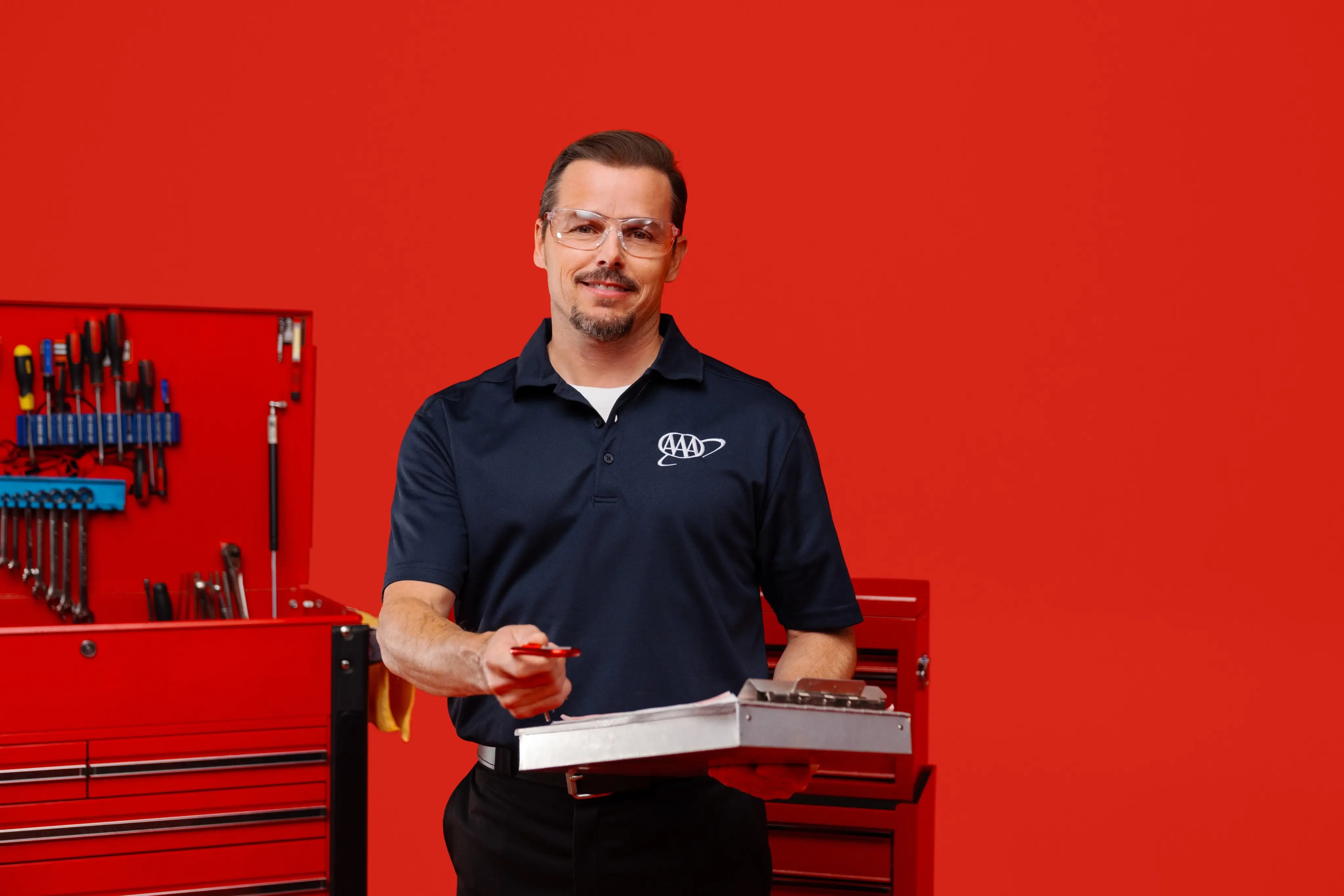 A male mechanic wearing safety glasses and a dark navy AAA polo shirt, holding a metal clipboard and pen while standing in front of a red tool chest against a solid red backdrop.
